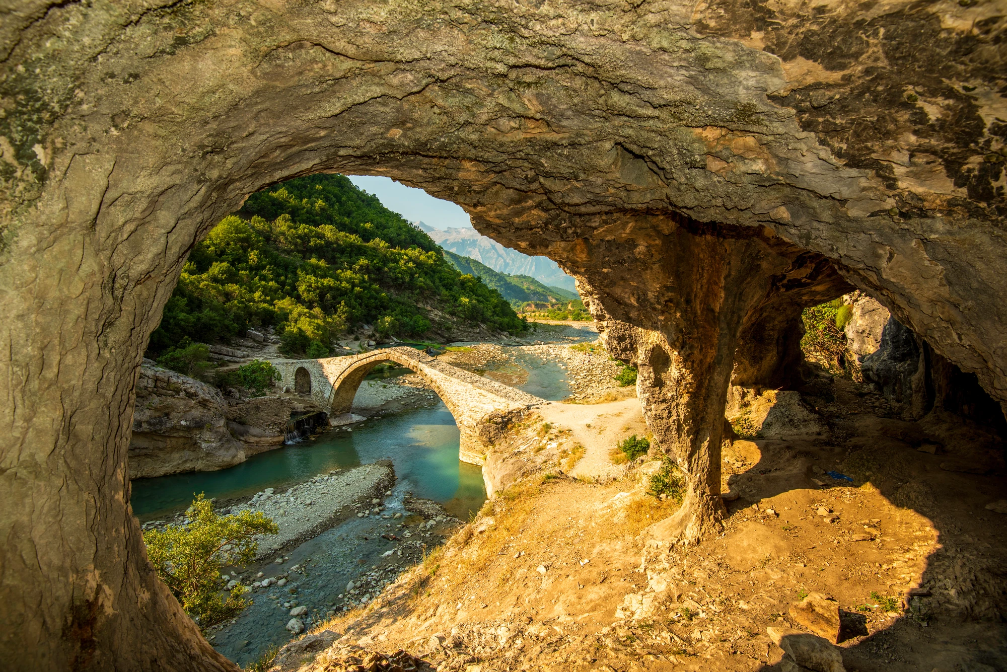 Albanien: Eine Kalksteinhöhle rahmt die Ura e Kadiut (Kadiu-Brücke) in Banjë Permet ein, wo ein steinerner Bogen den türkisfarbenen Fluss Vjosa in der üppigen albanischen Landschaft überspannt.