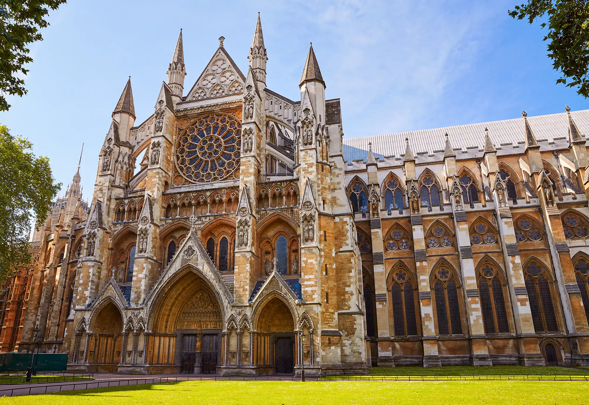 Das Bild zeigt die prächtige Westminster Abbey in London, eine der bedeutendsten historischen Kirchen Englands. Die gotische Architektur ist durch ihre spitzbogenförmigen Fenster und kunstvoll gearbeiteten Verzierungen besonders markant. Westminster Abbey ist bekannt als Ort königlicher Krönungen, Hochzeiten und Beerdigungen. Die Fassade zeigt die beeindruckenden Türme und das große Rosenfenster, die charakteristisch für gotische Kathedralen sind. Im Hintergrund sieht man den Turm der St Margaret Church, die neben der Westminster Abbey steht. Zusammen bilden diese beiden Gebäude ein ikonisches Wahrzeichen im Herzen Londons.
