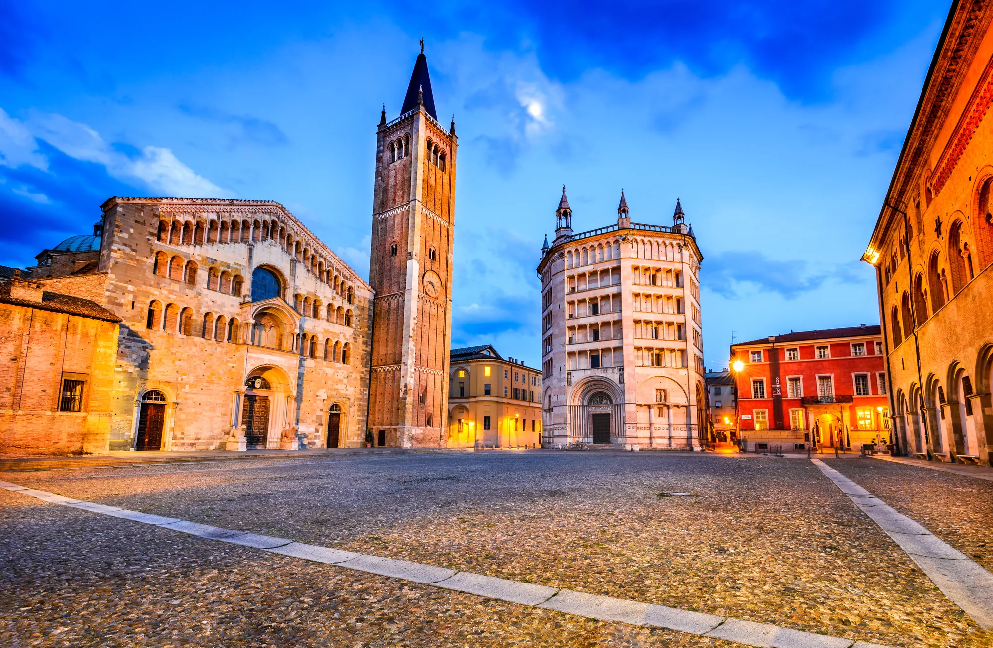 Der Domplatz in Parma bei Abenddämmerung, beleuchtet von stimmungsvoller Stadtbeleuchtung. Die prächtigen Gebäude und der imposante Dom erstrahlen in warmem Licht, wodurch eine einladende und romantische Atmosphäre entsteht.