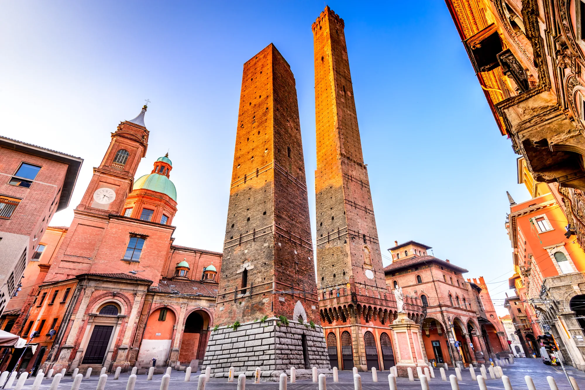 Vertikale Aufnahme der Geschwistertürme Asinelli und Garisenda, die majestätisch über der Stadt Bologna aufragen. Umgeben von historischen Gebäuden, erstrahlen die Türme bei sonnigem Wetter, während die warme Sonne die Architektur der Altstadt betont.
