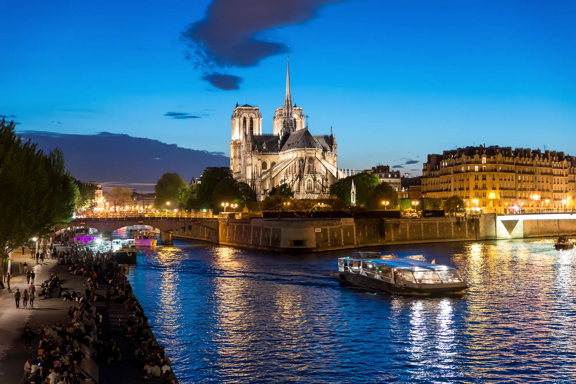 Panoramablick vom Flussufer auf die Seine mit beleuchteten Schifffahrten. Auf dem gegenüberliegenden Ufer ragt die beleuchtete Notre-Dame hervor, umgeben von Stadtbeleuchtung, die sich auf der Wasseroberfläche spiegelt. Am Ufer genießen zahlreiche Besucher die atmosphärische Abendstimmung.