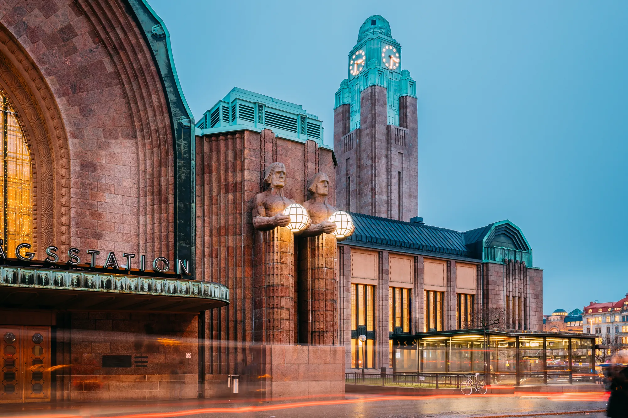 Helsinki Hauptbahnhof mit zwei großen Statuen, die eine Lichtkugel halten, und einer Fassade aus finnischem Granit. Der markante Uhrturm und die Statuen von Emil Wikström gelten als Wahrzeichen von Helsinki.