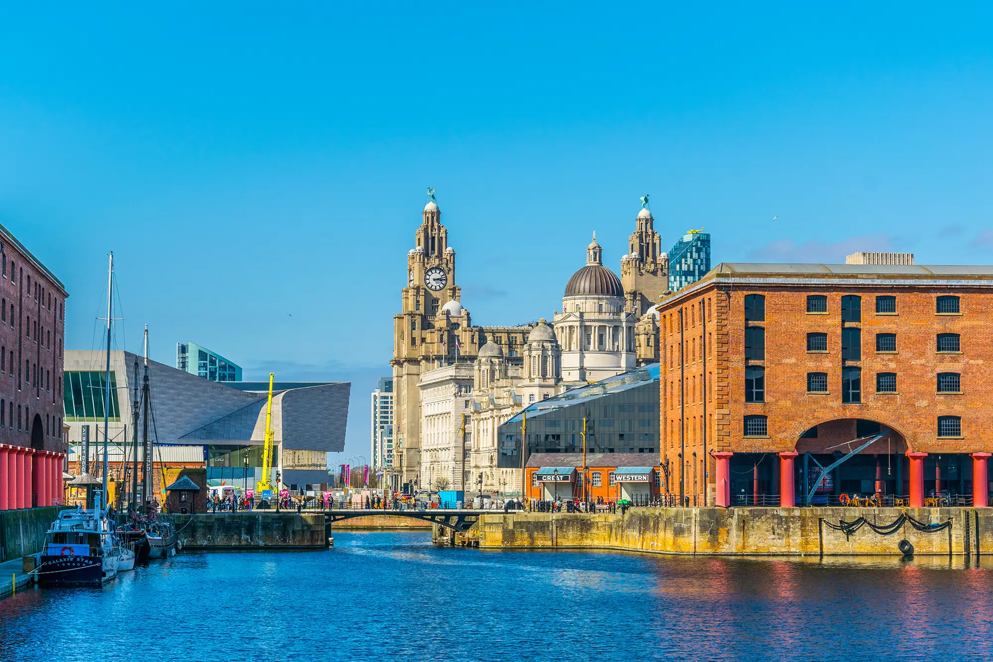 Das Bild zeigt den historischen Albert Dock in Liverpool, der für seine beeindruckende Architektur und seine kulturelle Bedeutung bekannt ist. Im Vordergrund fließt das ruhige Wasser des Hafens, gesäumt von traditionellen roten Backsteingebäuden. Der ikonische Royal Liver Building mit seinen charakteristischen Türmen und dem Clock Tower erhebt sich majestätisch im Hintergrund, während moderne Bauwerke harmonisch in die Szene integriert sind. Die lebendige Atmosphäre wird durch die Menschenmengen und die Boote im Hafen verstärkt, was diesen Ort zu einem beliebten Ziel für Touristen und Einheimische macht.