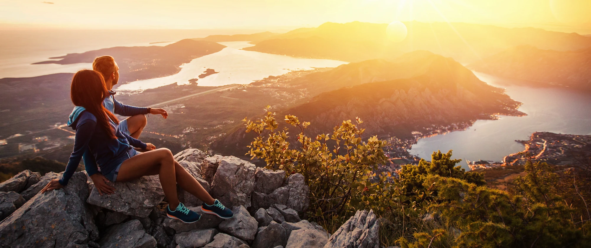 Zwei Personen sitzen auf einem Felsen und blicken auf eine weite Landschaft mit Bergen und einem Gewässer bei Sonnenuntergang.