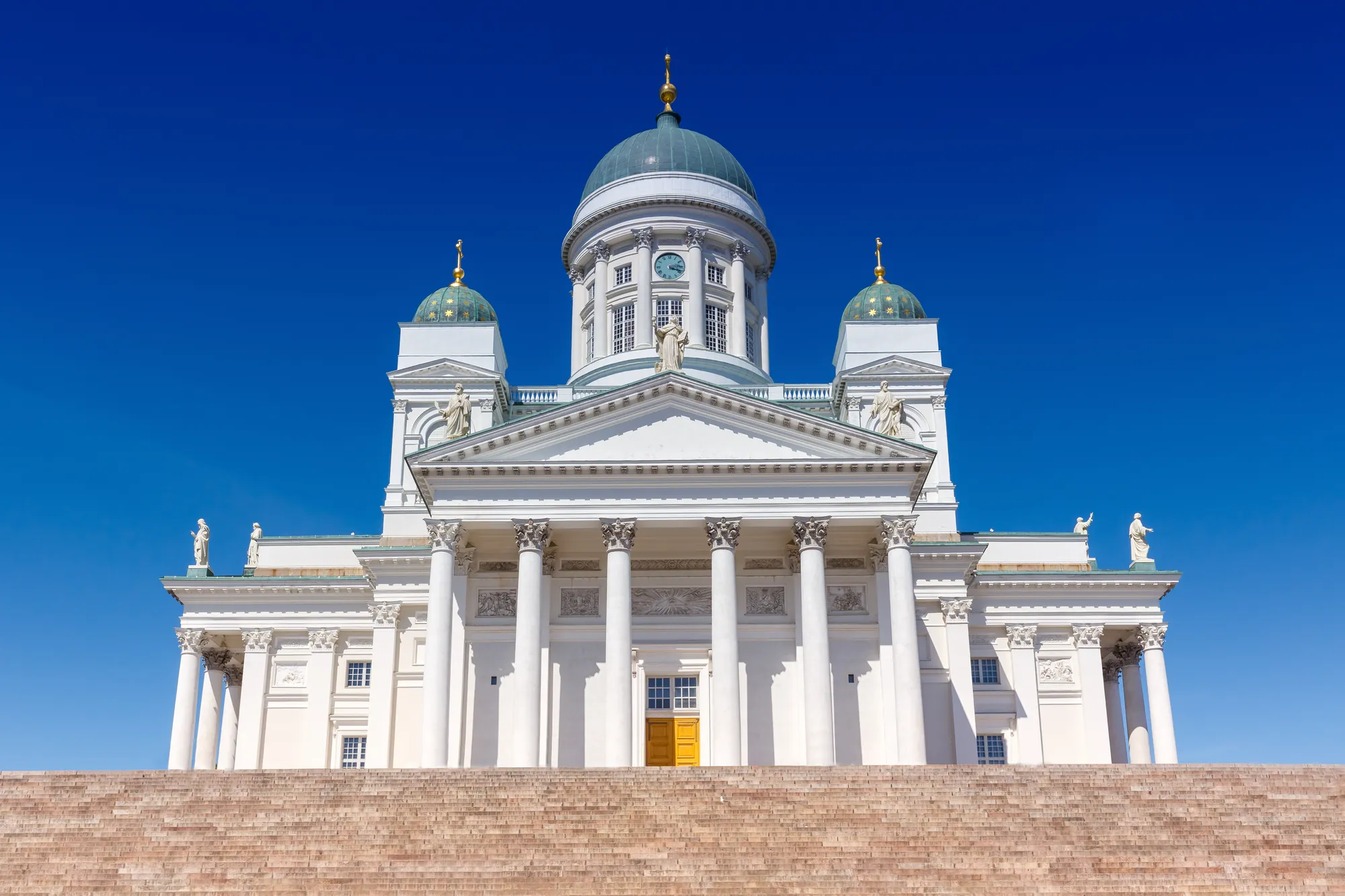 Frontansicht der Dom-Kathedrale von Helsinki, von unten fotografiert mit den breiten Treppen im Vordergrund. Das weiße, majestätische Gebäude erstrahlt bei sonnigem und klarem Wetter eindrucksvoll im Licht.