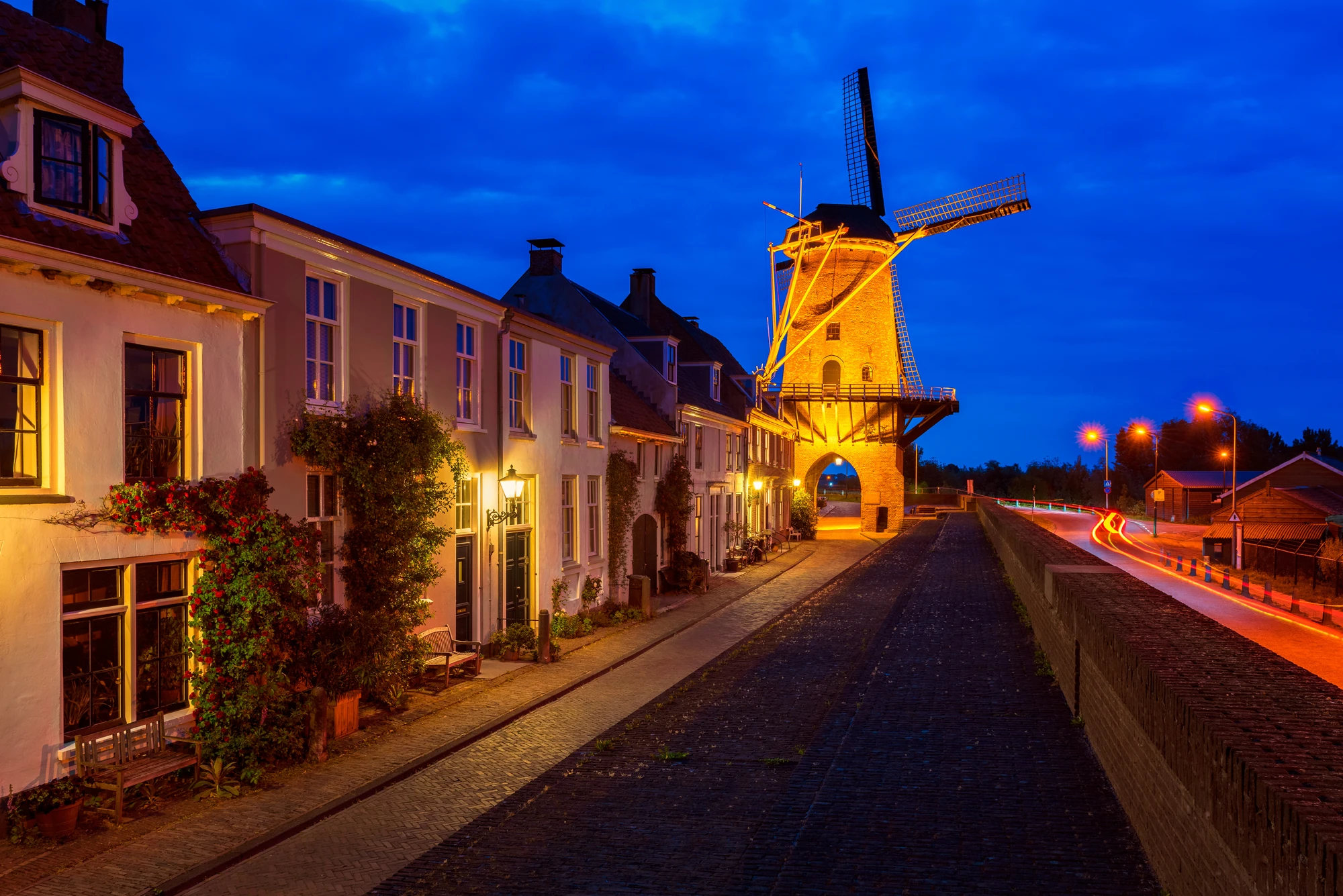 Beleuchtete Windmühle am Ende einer gepflasterten Straße mit Reihenhäusern und Straßenlaternen bei Abenddämmerung. Wijk bij Duurstede