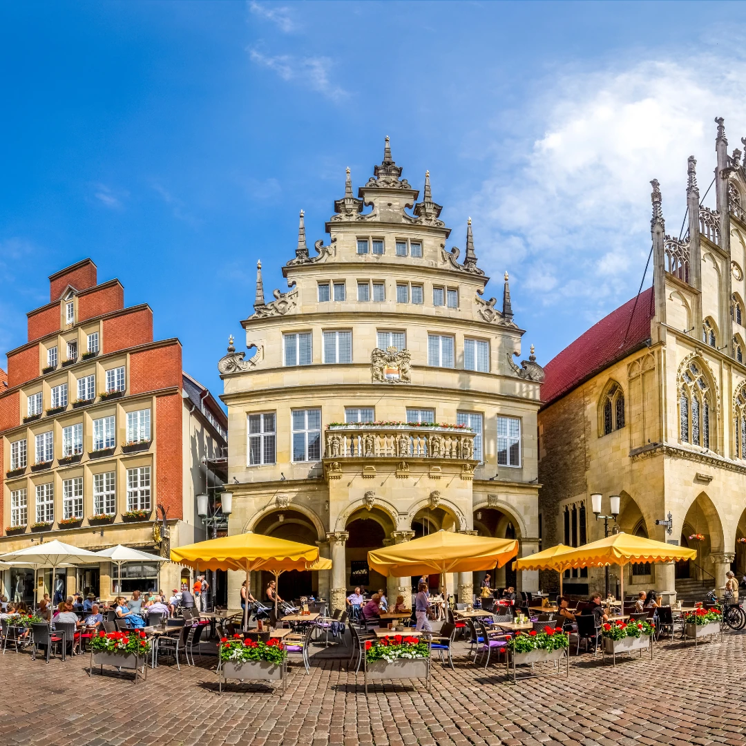 Das Bild zeigt eine lebendige Szene aus der Altstadt von Münster. Im Vordergrund befindet sich ein Marktplatz mit gemütlichen Straßencafés, gelben Sonnenschirmen und blühenden Blumenarrangements. Umgeben wird der Platz von historischen Gebäuden mit prachtvollen Fassaden, darunter das imposante Rathaus von Münster mit seiner gotischen Architektur. Der Himmel ist blau mit einigen weißen Wolken, was der Szene eine freundliche und einladende Atmosphäre verleiht.