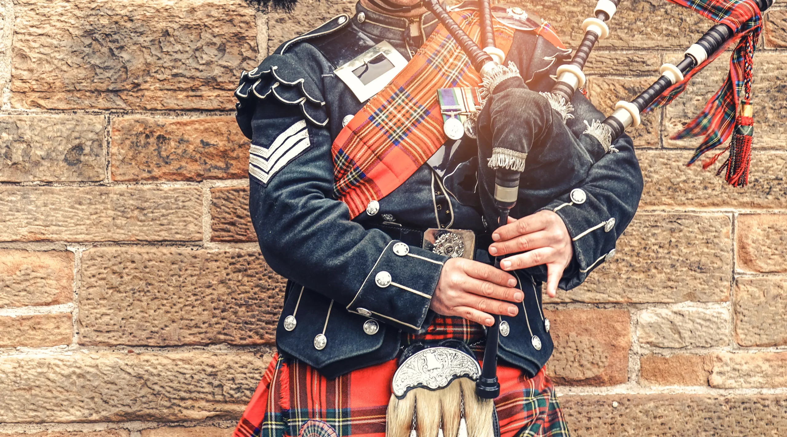 Schottischer Dudelsackspieler in traditionellem rot-schwarzen Tartan-Outfit steht vor einer Steinmauer. Edinburgh, das beliebteste Reiseziel in Schottland.