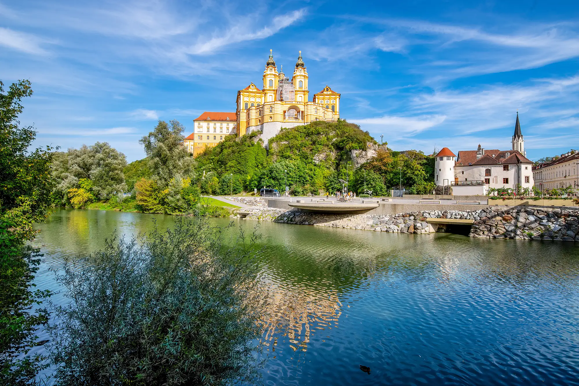 Stift Melk in der Wachau mit Stadt und Gebäuden im Vordergrund am Ufer der Donau mit Bäumen und Büschen