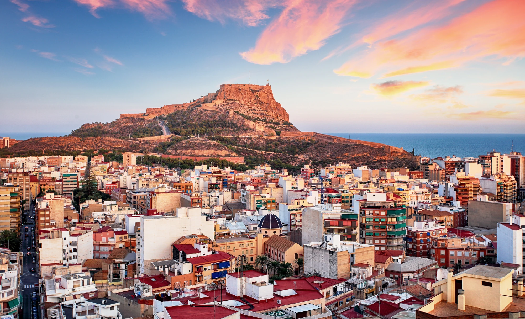 Alicante Blick auf eine Stadt mit vielen Gebäuden und einem großen Felsen mit Festung im Hintergrund bei Sonnenuntergang.