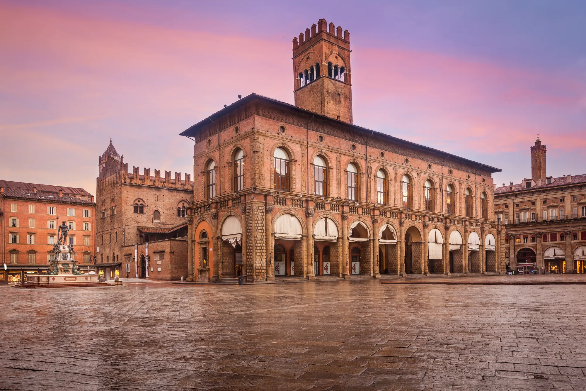 Aussicht auf den beeindruckenden Hauptplatz Piazza Maggiore in Bologna bei sanfter rosafarbener Abenddämmerung. Die historischen Gebäude und die majestätische Basilika werden in warmes Licht getaucht, wodurch eine romantische und stimmungsvolle Atmosphäre entsteht.