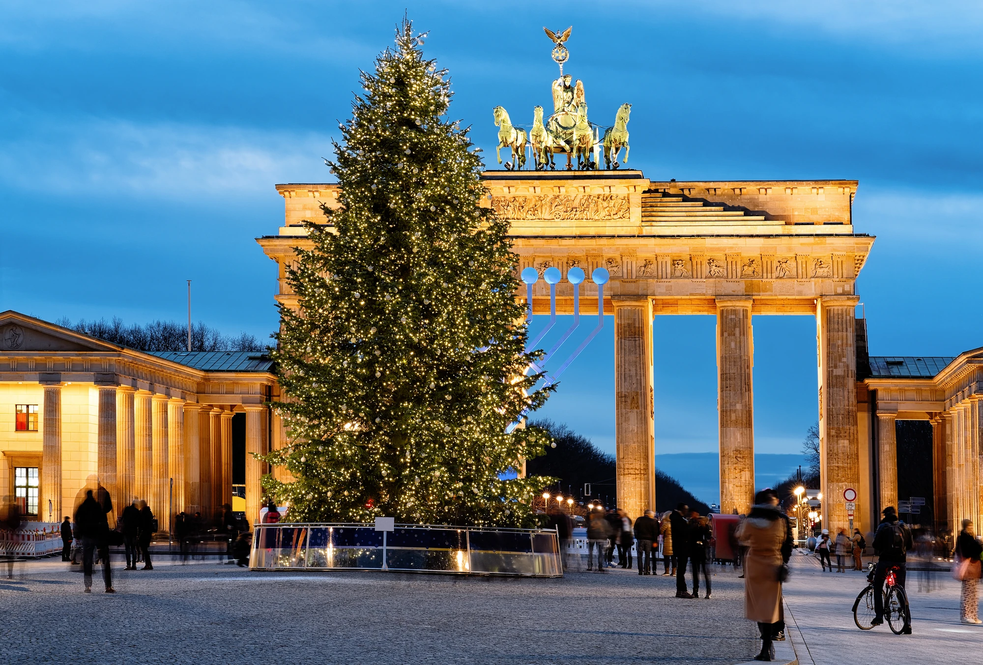 Beleuchteter Weihnachtsbaum vor dem Brandenburger Tor bei Dämmerung, Menschen im Vordergrund sichtbar.