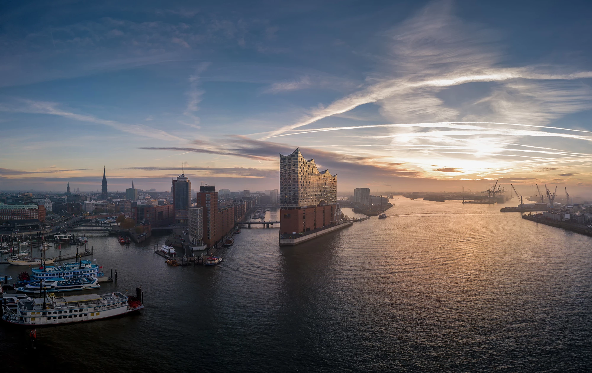Eine atemberaubende Luftaufnahme der Elbphilharmonie in Hamburg bei Sonnenuntergang, mit einem weiten Blick auf den Hafen und die umgebenden Wasserstraßen. Der Himmel ist von dramatischen Wolken und Sonnenstrahlen durchzogen, die moderne Architektur des Gebäudes in goldenes Licht tauchen.