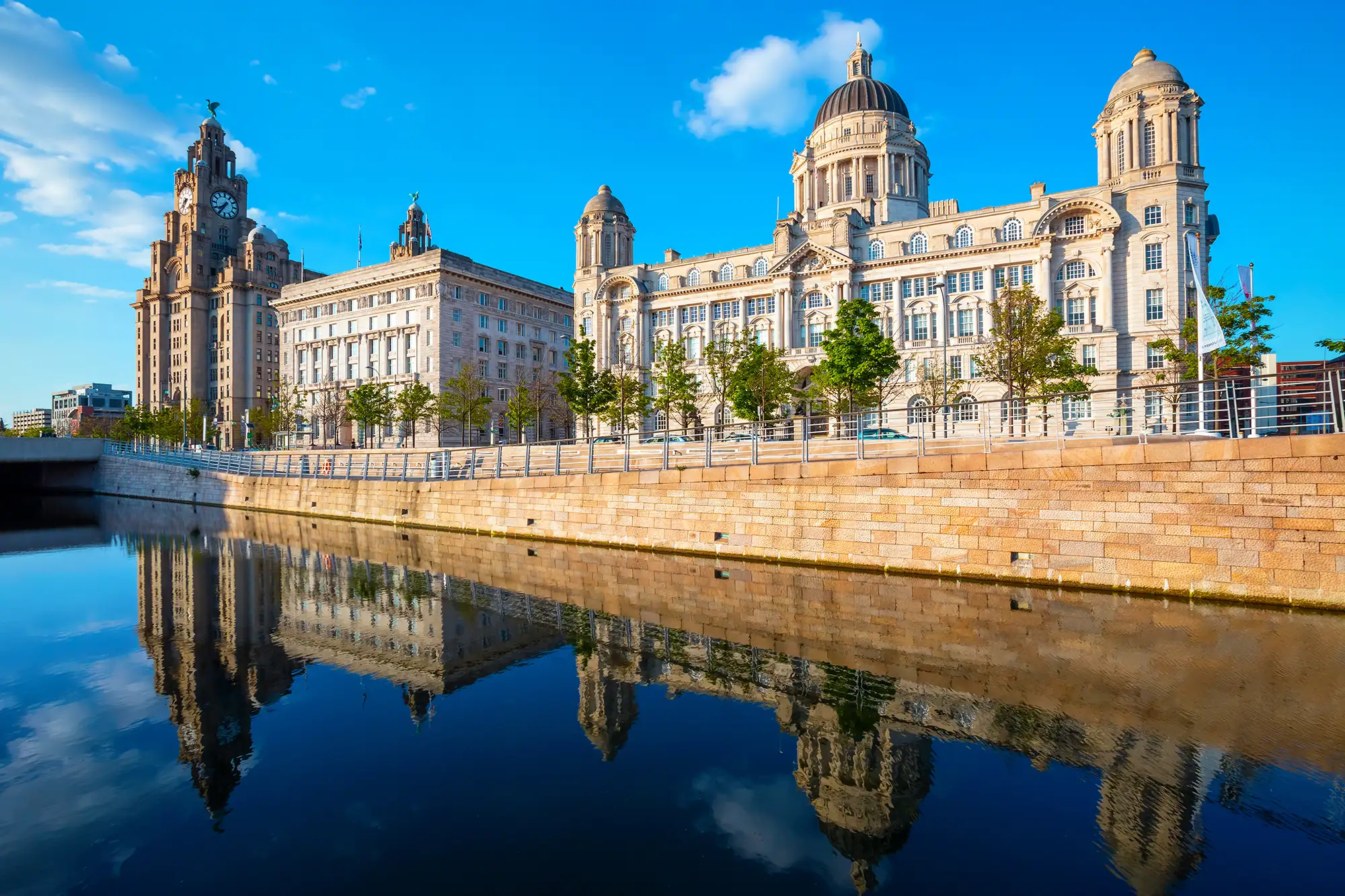 Das Bild zeigt die prächtige Architektur des Liverpool Piers, das für seine beeindruckenden Gebäude und die ruhige Wasserfläche bekannt ist. Im Vordergrund spiegelt sich die historische Skyline der Stadt im klaren Wasser wider, was eine malerische und harmonische Szenerie schafft. Die markanten Bauwerke, darunter der Royal Liver Building mit seiner charakteristischen Uhr und der Liver Bird, ragen majestätisch in den strahlend blauen Himmel. Diese Ansicht verkörpert den Charme und die kulturelle Bedeutung von Liverpool als Hafenstadt.