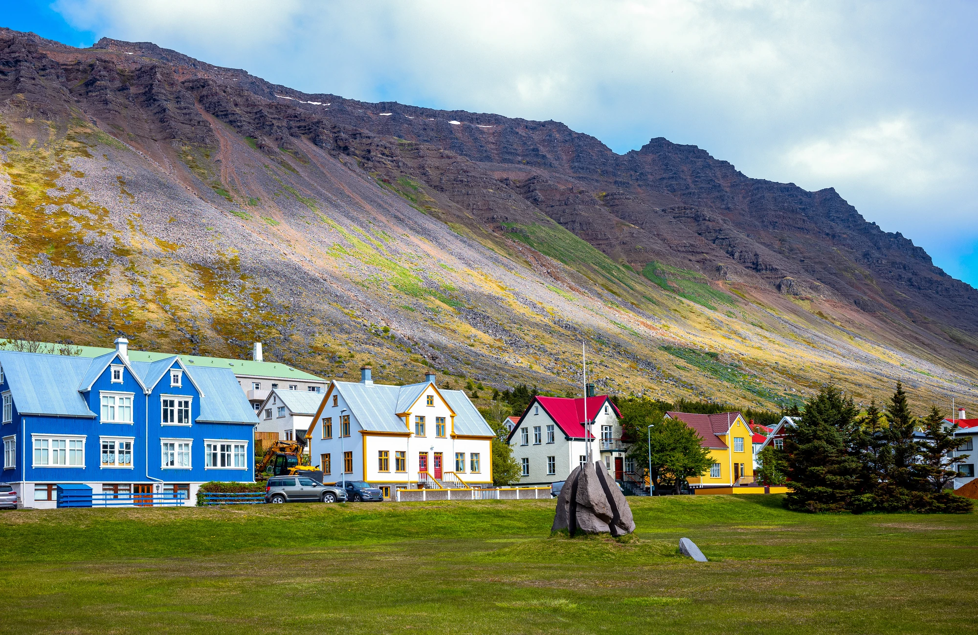 Ísafjörður (Island) Bunte Häuserreihe vor steilem, felsigem Berg mit grünem Wiesenfeld und großem Felsen im Vordergrund.