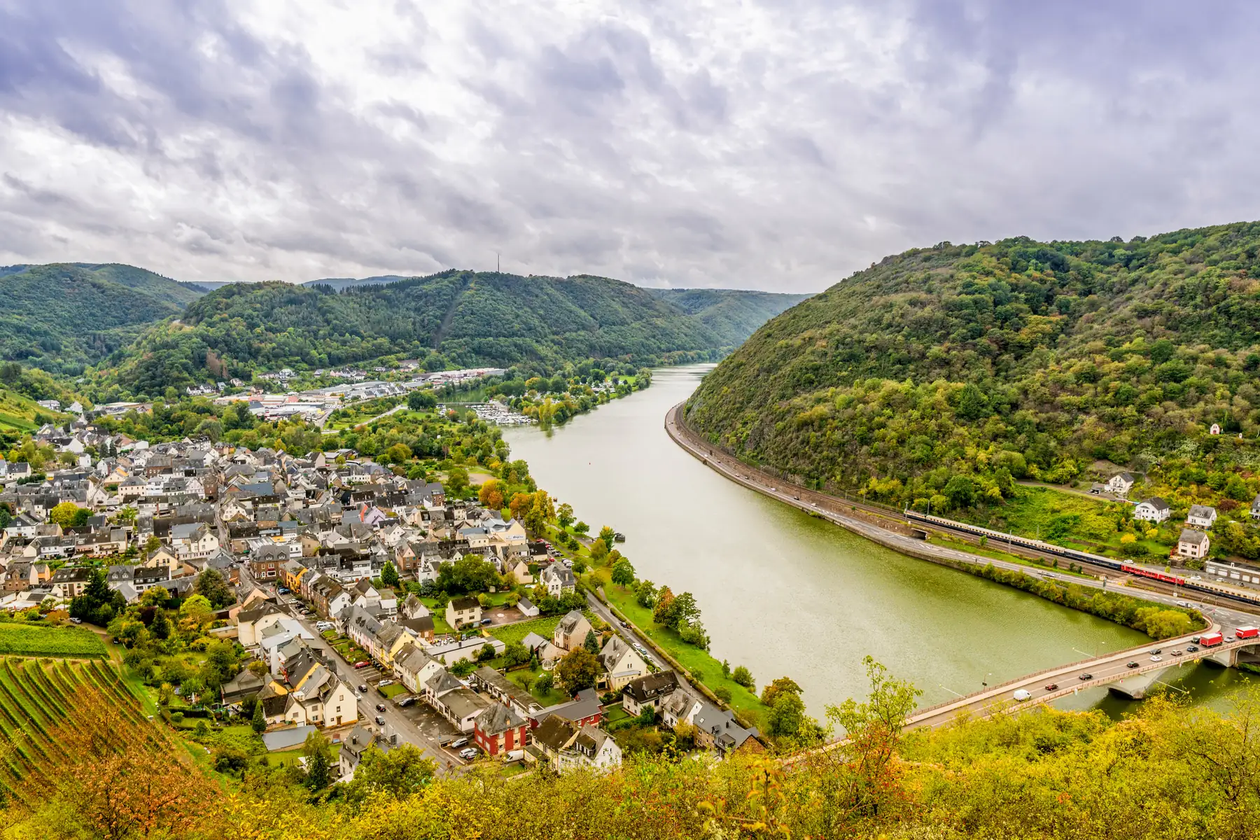 Blick auf die Mosel, die sich durch ein bewaldetes Tal mit einer Stadt (Tris-Karden) und einer Brücke schlängelt