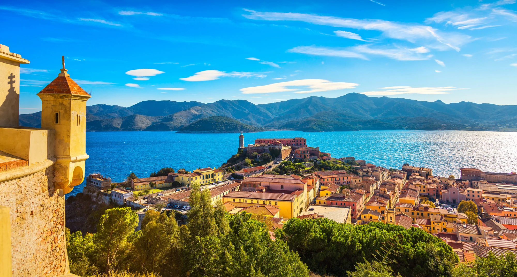 Elba Blick auf eine Küstenstadt mit roten Dächern, einem Leuchtturm auf einer Landzunge und Bergen im Hintergrund unter blauem Himmel.