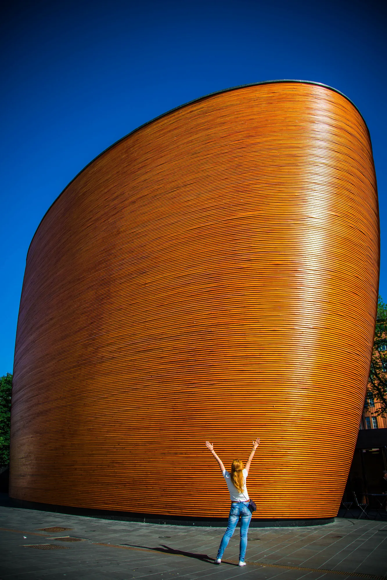 Frau mit hoch ausgestreckten Armen vor der Kamppi-Kapelle, einem braunen gewölbten Gebäude, das durch seine moderne Architektur und einladende Atmosphäre besticht.