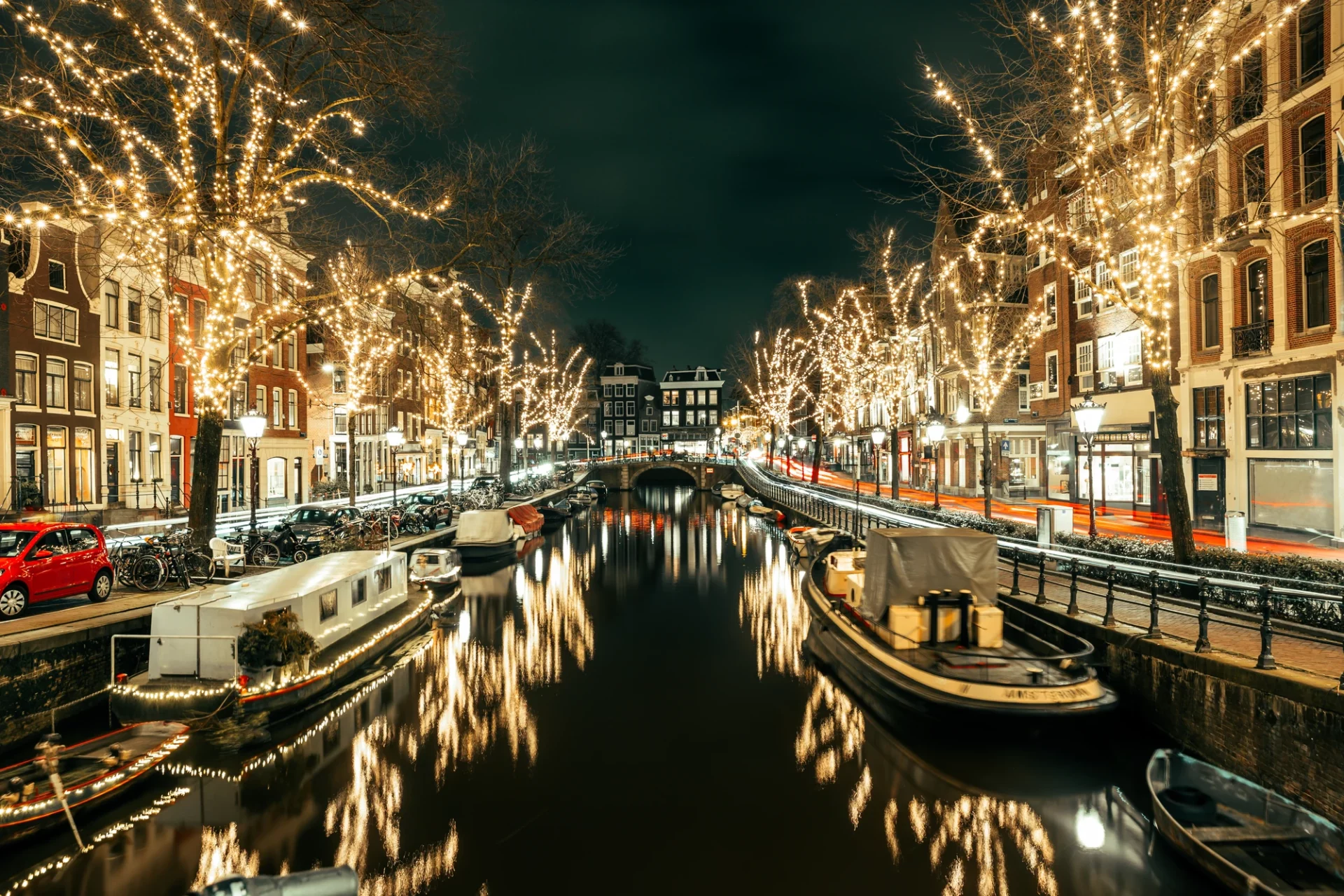 Blick auf die Amsterdamer Gracht bei Nacht, wo die Weihnachtsbeleuchtung an den Bäumen und die Stadtbeleuchtung eine festliche Stimmung erzeugen. Die Lichter spiegeln sich sanft auf der Wasseroberfläche des Kanals und verleihen der Szene einen magischen Glanz.
