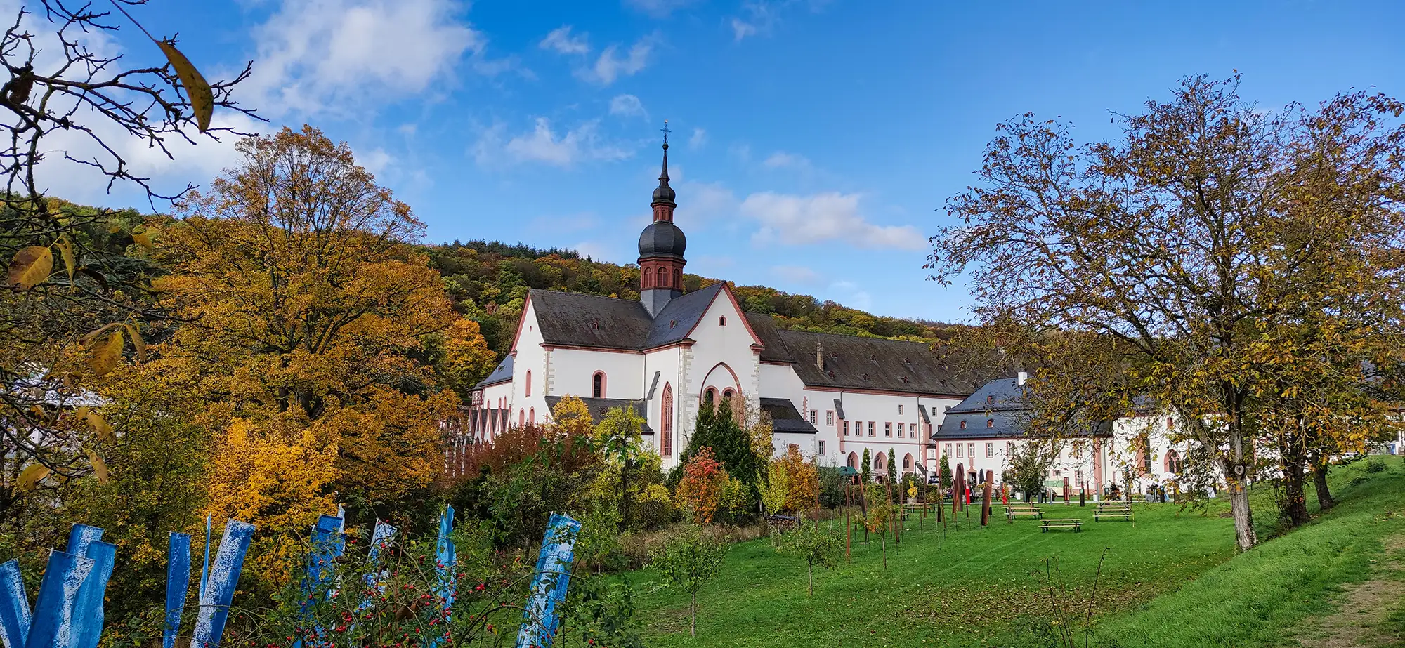 Kloster Eberbach in Eltville am Rhein im Rheingau mit Bäumen und Rasen mit Bänken und Büschen vor einer Bergkette im Herbst