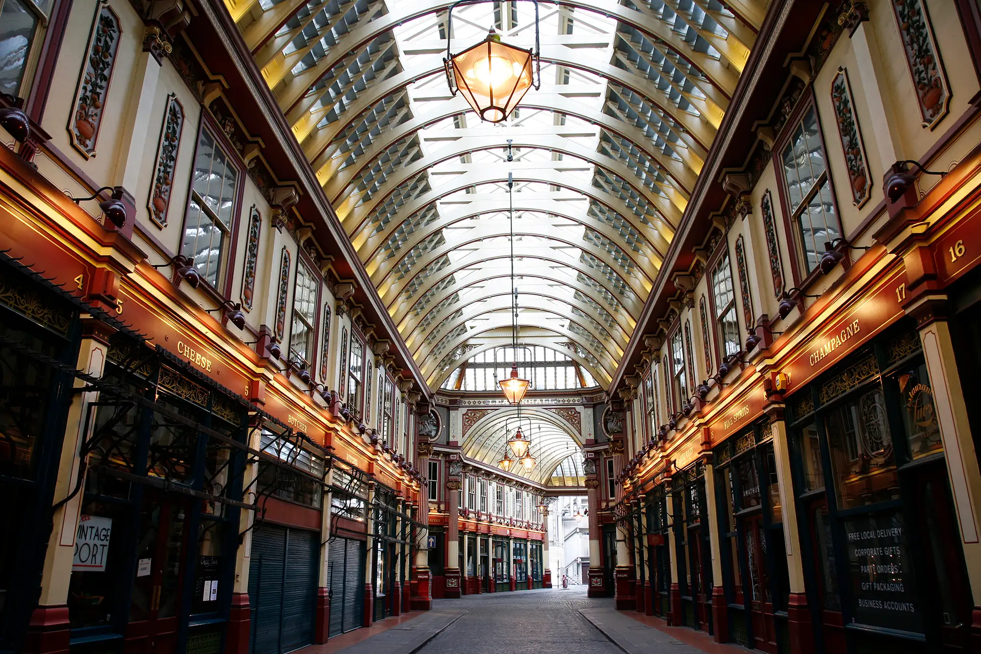 Das Bild zeigt die Leadenhall Market, eine historische überdachte Markthalle in London. Der Markt ist bekannt für seine aufwendige viktorianische Architektur mit verzierten Fassaden, eleganten Bögen und einem atemberaubenden Glasdach, das viel natürliches Licht in die Passage lässt. Die Geschäfte entlang der Seiten sind mit kunstvollen Schildern und detaillierten Dekorationen versehen, die eine warme und einladende Atmosphäre schaffen. Die Lampen, die von der Decke hängen, verstärken das elegante Ambiente dieses ikonischen Londoner Marktes.