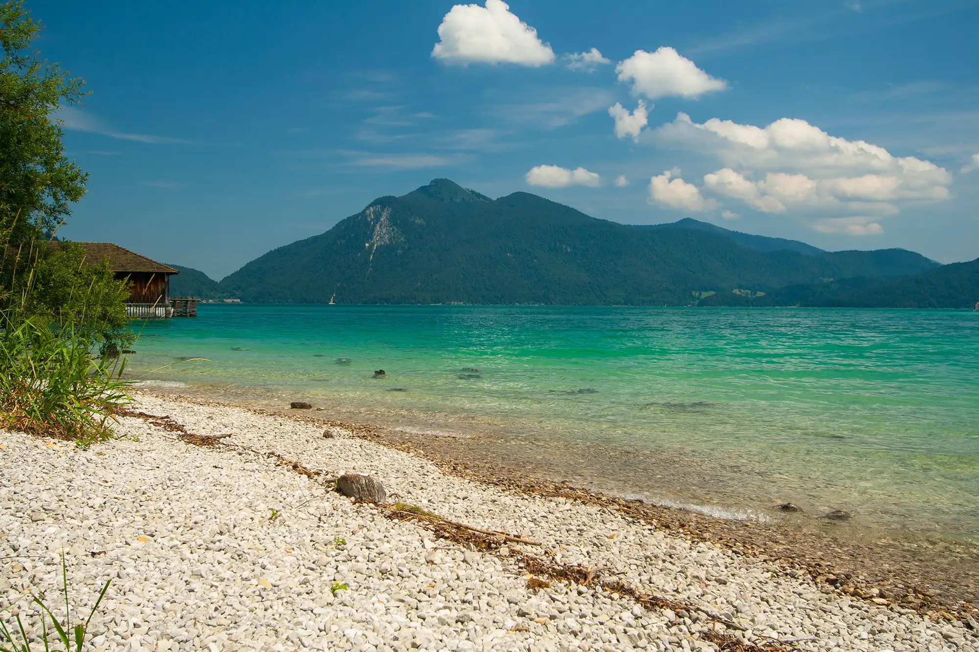 Kiesstrand am Walchensee in Bayern mit einer Holzhütte und Bergen mit Wald im Hintergrund