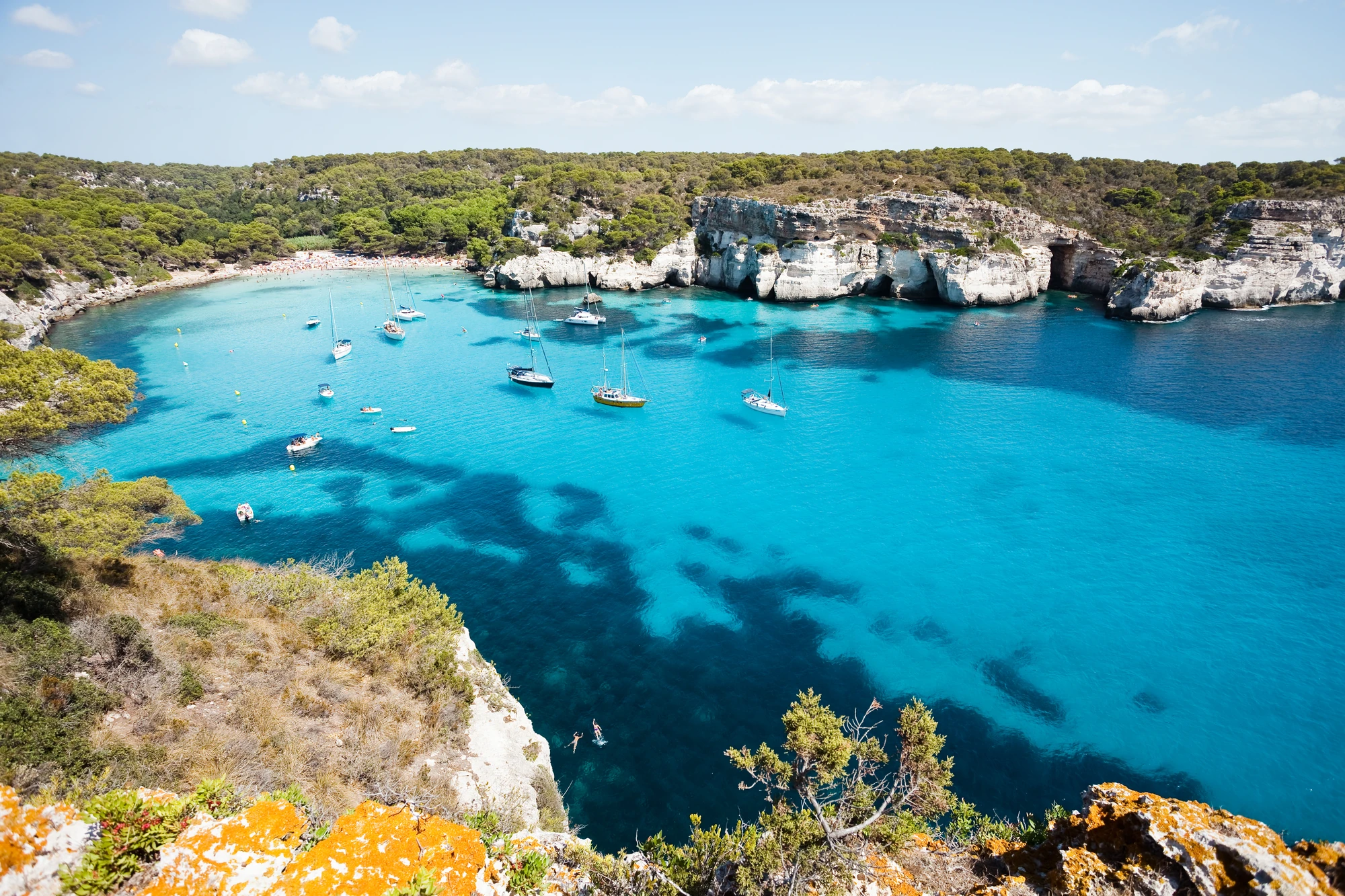 Menorca Bucht mit türkisblauem Wasser, umgeben von bewaldeten Klippen, mehrere Segelboote auf dem Wasser.