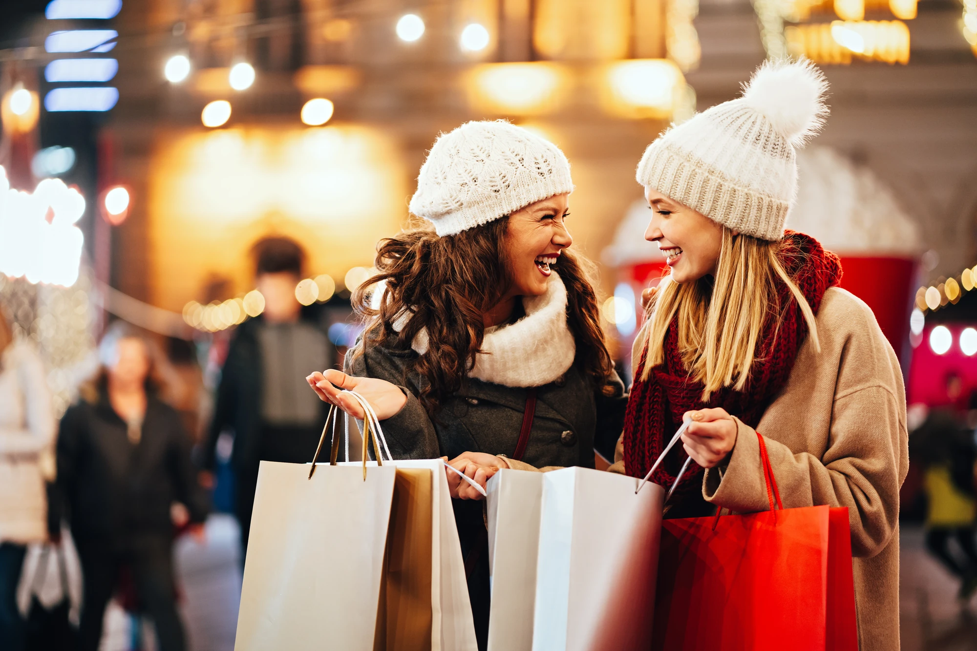 Das Bild zeigt zwei Frauen, die in einer festlich beleuchteten Stadt einkaufen. Sie tragen warme Winterkleidung und Mützen, während sie lachend ihre Einkaufstüten präsentieren. Die Szene vermittelt eine fröhliche und weihnachtliche Atmosphäre, unterstützt von den hellen Lichtern im Hintergrund.