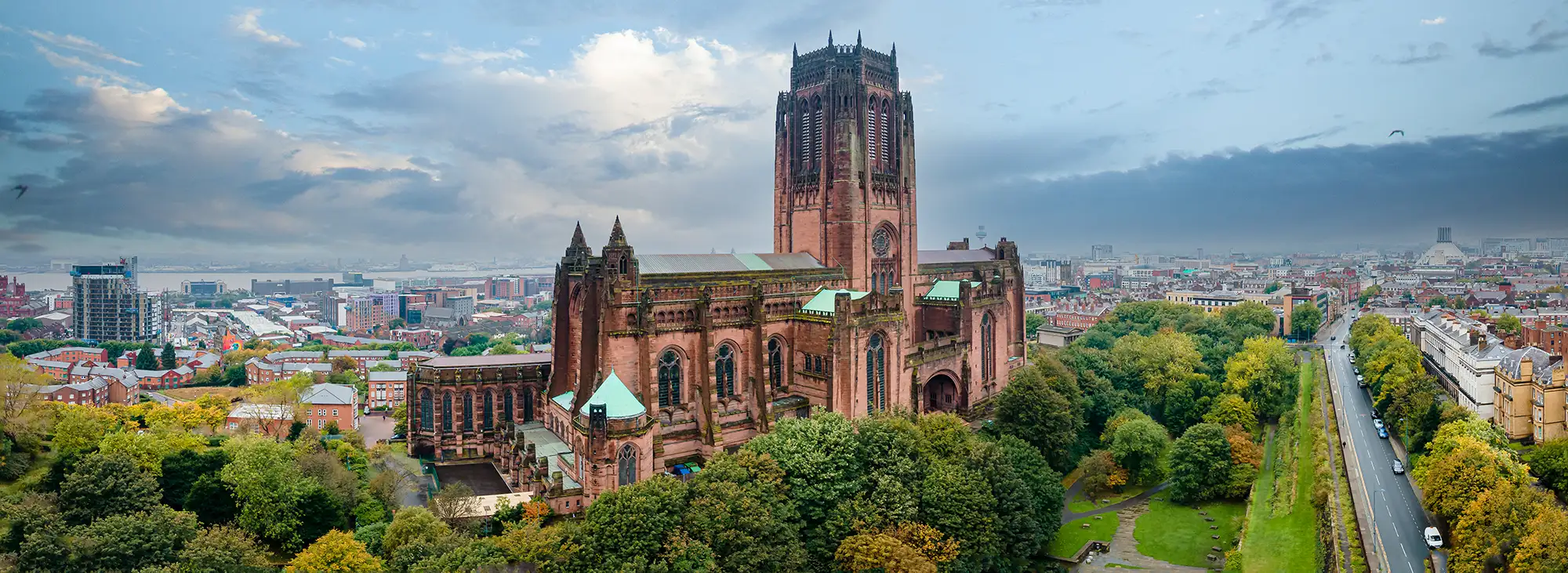 Das Bild zeigt die beeindruckende Liverpool Cathedral, die für ihre markante gotische Architektur bekannt ist. Die Kathedrale, die als größte Kirche Großbritanniens gilt, erhebt sich majestätisch über die Stadt und ist umgeben von einer üppigen grünen Landschaft. Im Hintergrund sind moderne Stadtstrukturen und der Hafen von Liverpool sichtbar, was den Kontrast zwischen Tradition und Moderne unterstreicht. Der Himmel ist teilweise bewölkt, was eine dramatische Stimmung schafft und die monumentale Präsenz der Kathedrale hervorhebt.