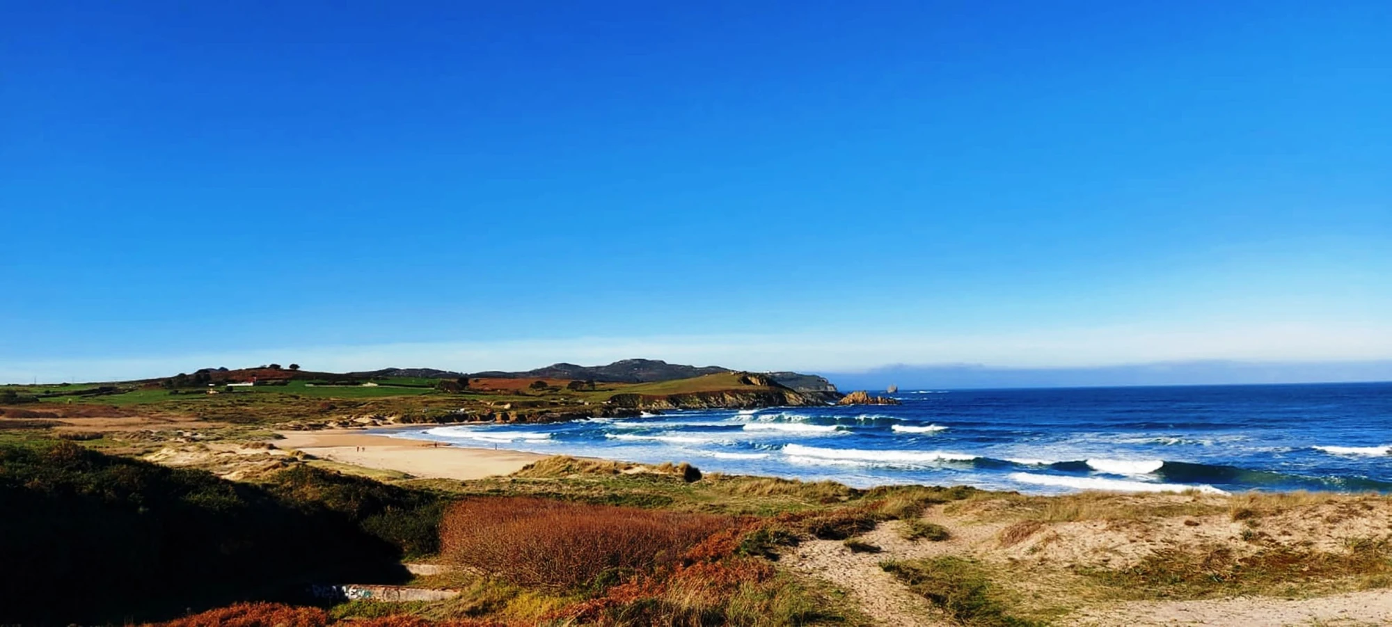Ferrol Küstenlandschaft mit Sandstrand, Dünen und Wellen unter klarem blauem Himmel.