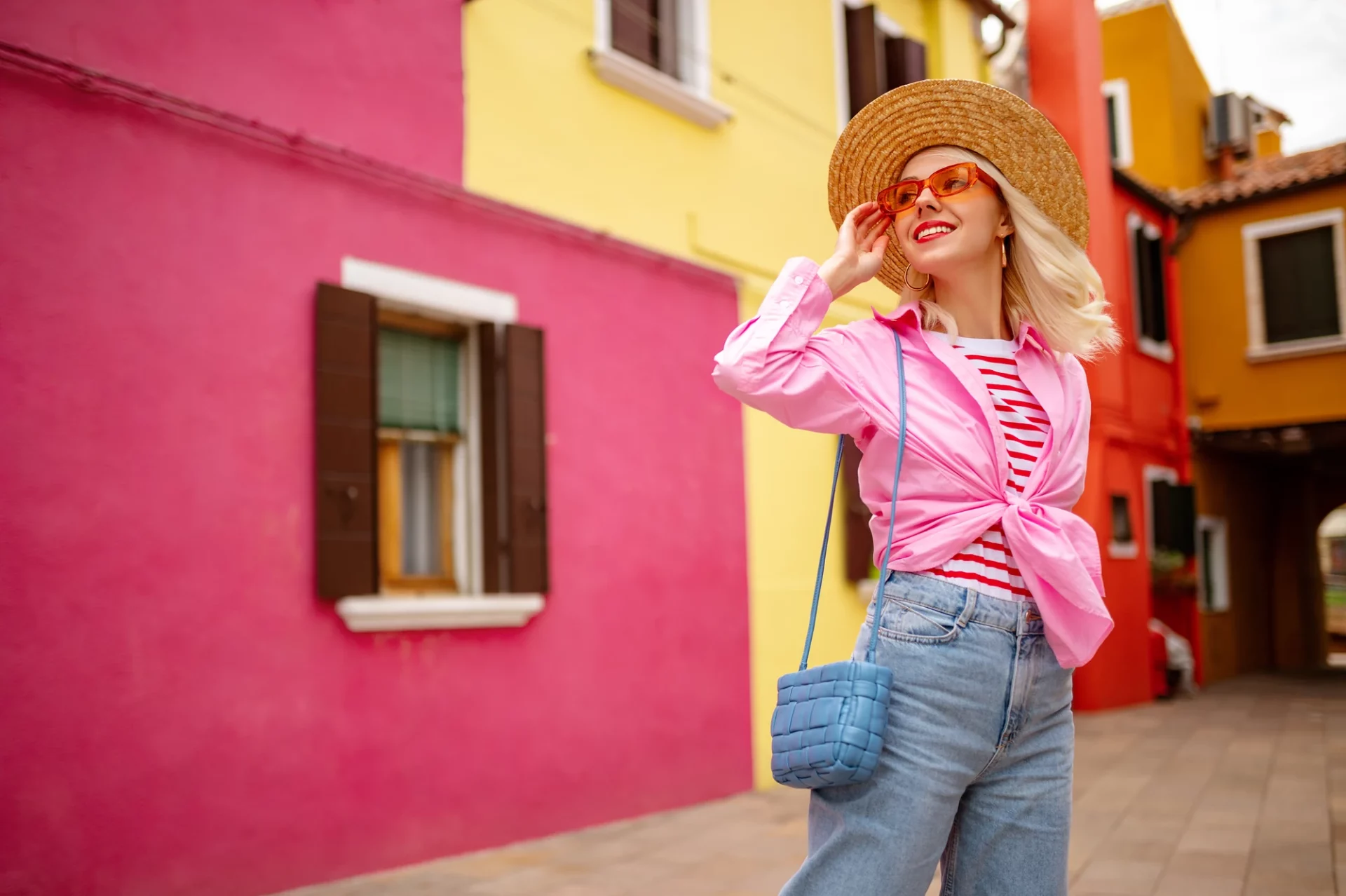 Eine Frau in strahlenden Farben steht in der farbenfrohen Stadt Burano, umgeben von den leuchtenden, bunten Häusern. Das Tageslicht verstärkt die lebendige Atmosphäre und hebt die Schönheit der Architektur und der Umgebung hervor, während die Frau harmonisch in die lebendige Szenerie integriert ist.
