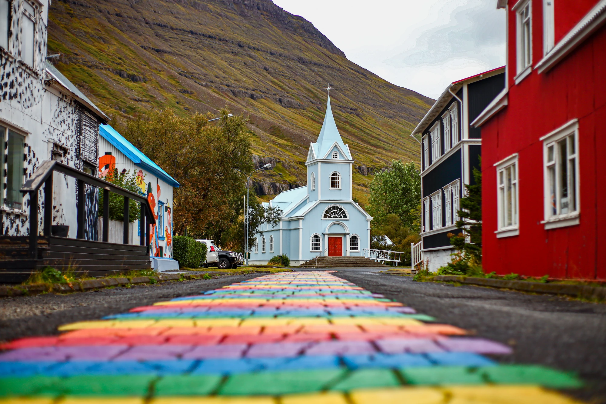 Seyðisfjörður (Island) Bunte gepflasterte Straße führt zu einer hellblauen Kirche mit spitzem Turm, umgeben von bunten Häusern und Bergen im Hintergrund.