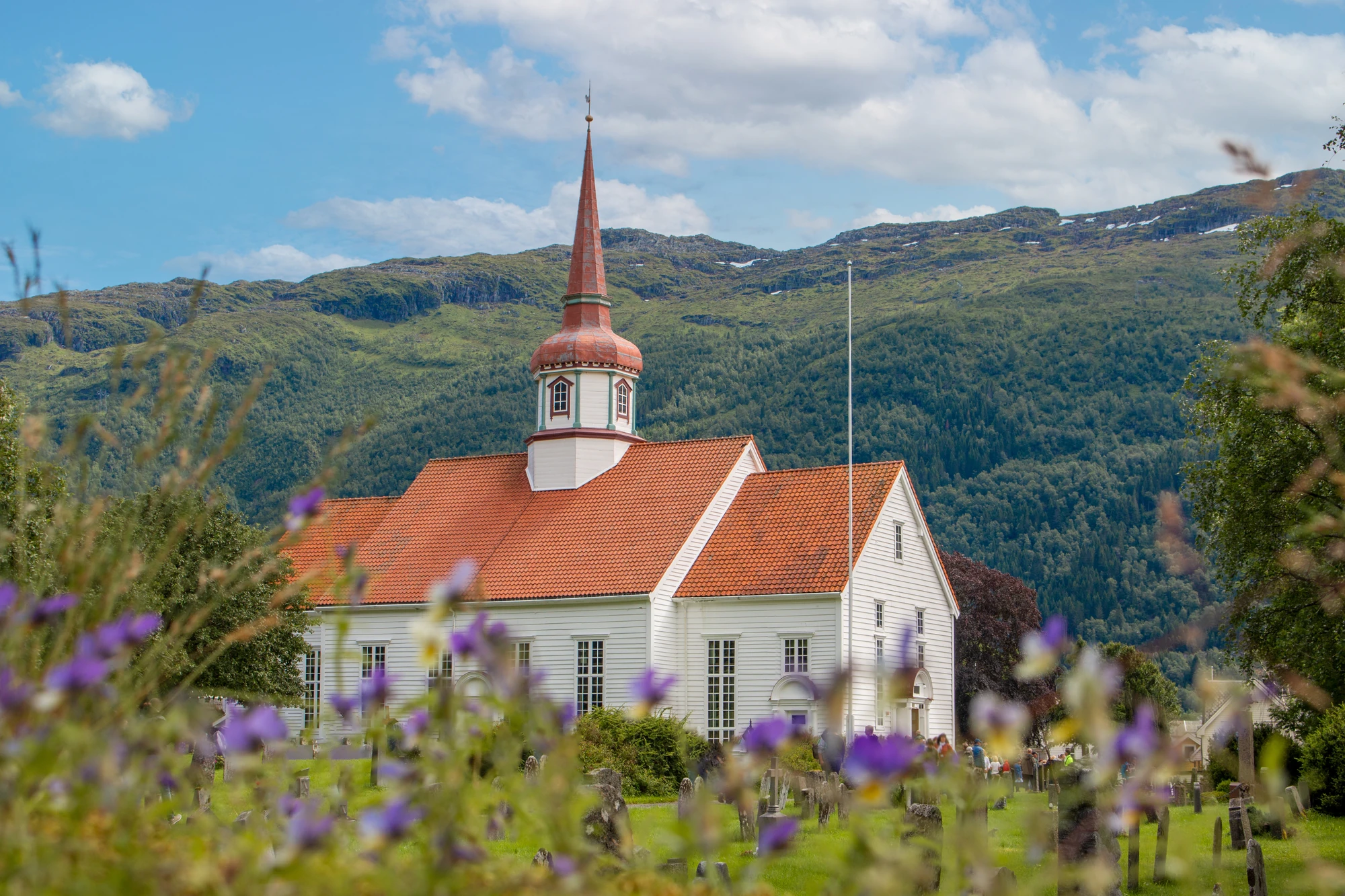 Weißes Kirchengebäude mit rotem Dach und spitzem Turm, umgeben von grünen Bergen und bunten Blumen im Vordergrund.