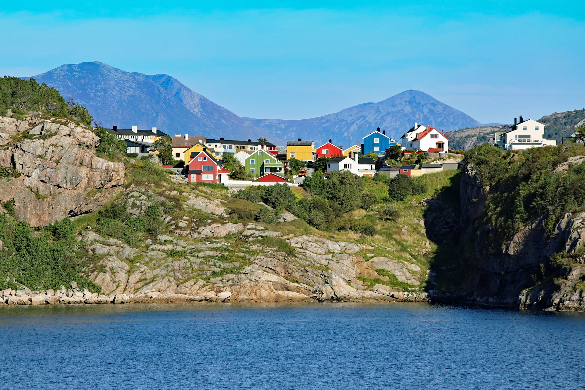 Bunte Häuser in verschiedenen Farben auf einem Hügel, umgeben von Bergen und Wasser. Klare Himmel mit wenigen Wolken.