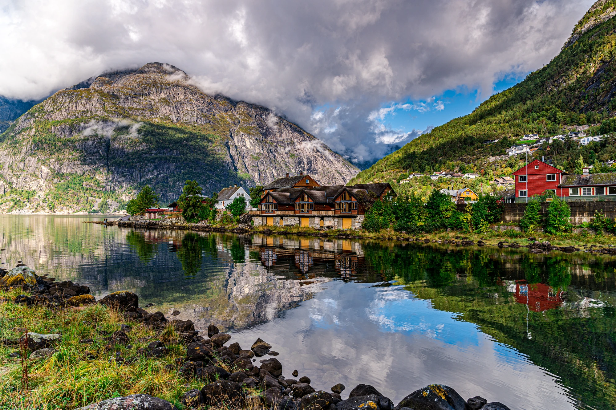 Berglandschaft mit See, mehreren Holzhäusern und bewölktem Himmel, Spiegelung im Wasser sichtbar.