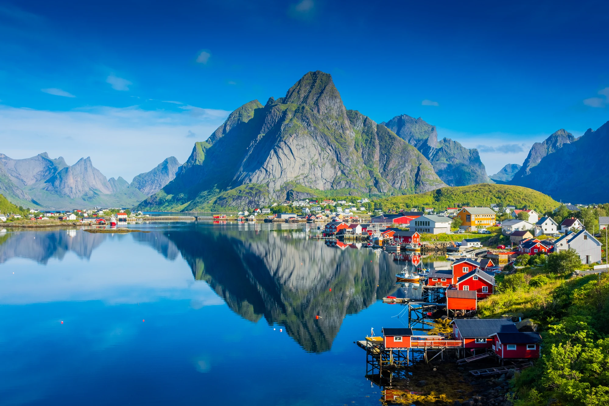 Lofoten Inseln Blick auf eine malerische Küstenstadt mit roten Häusern am Wasser, umgeben von Bergen und klarem Himmel, reflektiert im ruhigen Wasser.