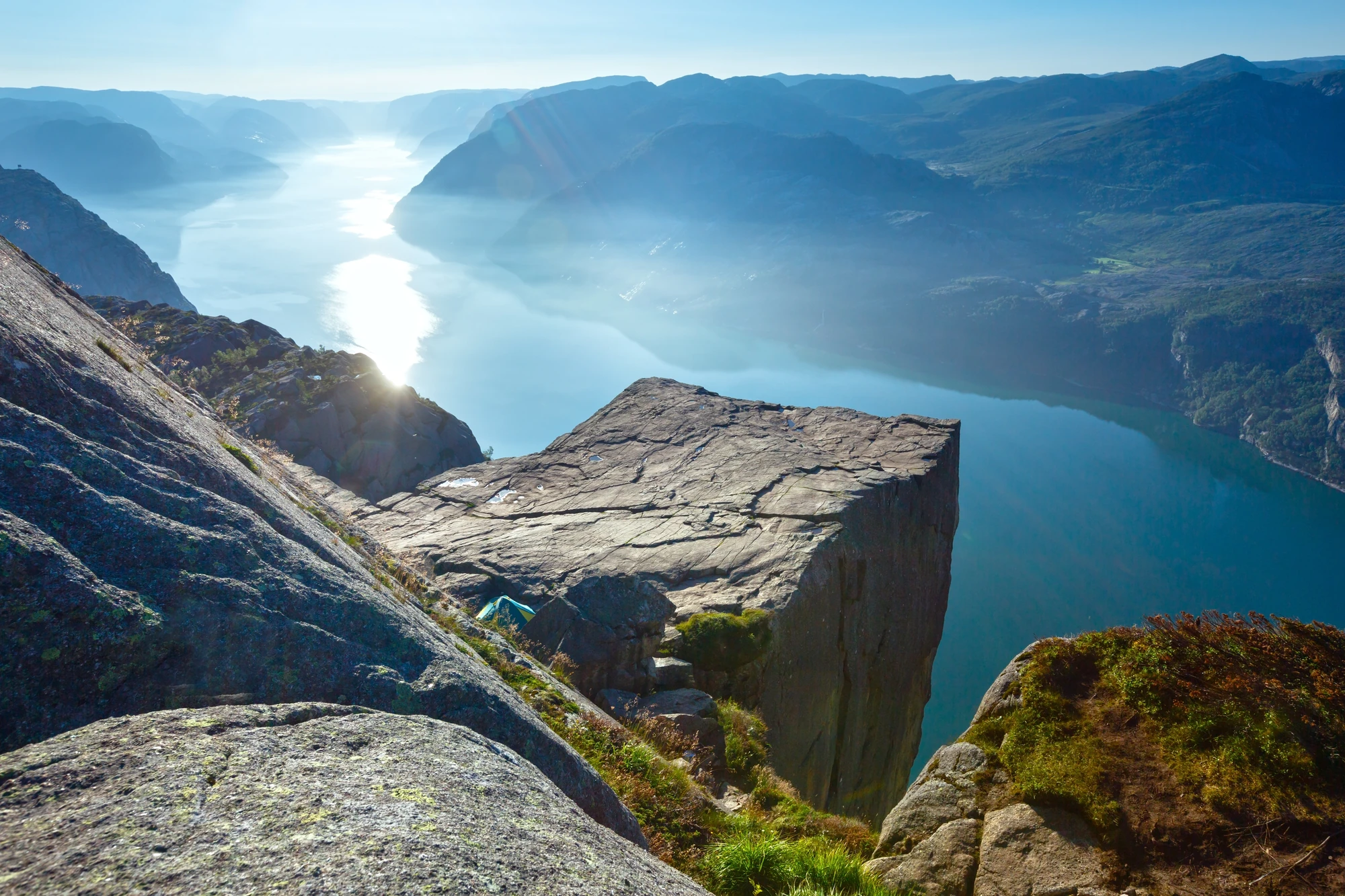 Felsplateau ragt über Fjord mit Bergen im Hintergrund, Sonnenlicht spiegelt sich auf dem Wasser.