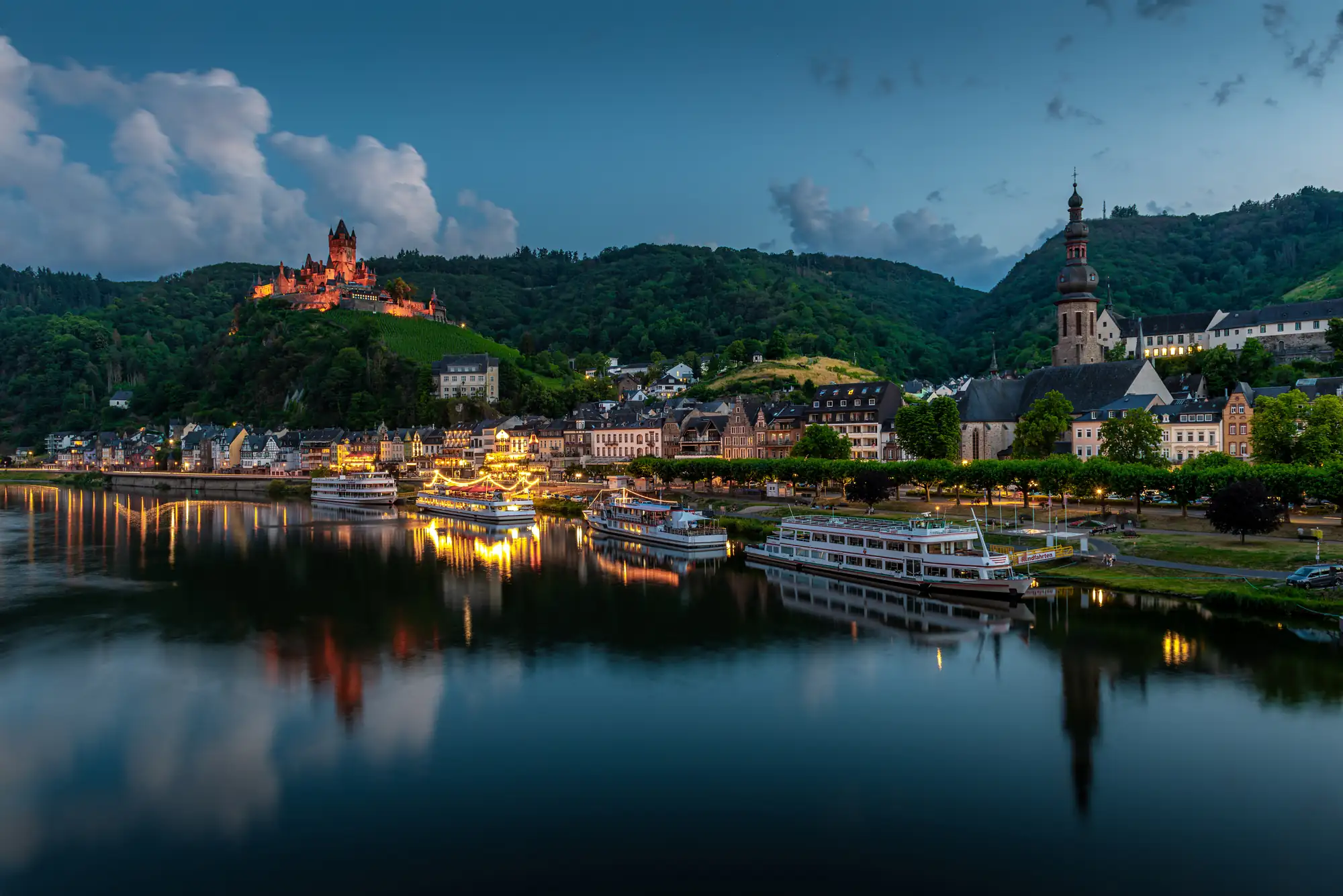 Abendliche Stadtansicht von Cochem an der Mosel mit beleuchtetem Reichsburg Cochem auf dem Hügel, Fluss und mehreren angelegten Schiffen