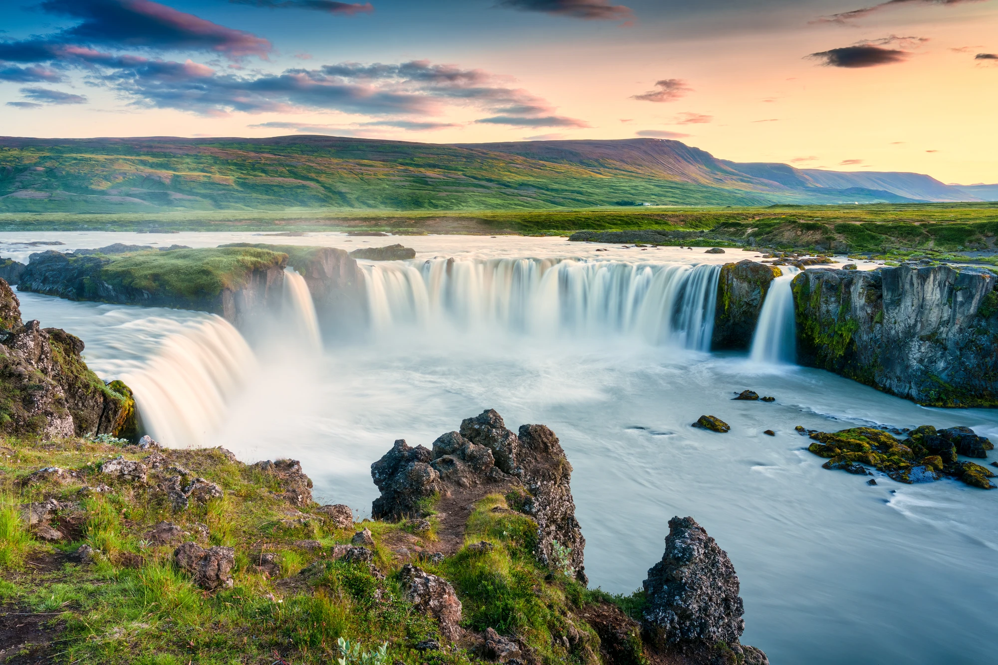 Akureyri (Island) Breiter Wasserfall mit mehreren Kaskaden, umgeben von grünen Wiesen und felsigem Gelände unter einem bewölkten Himmel bei Sonnenuntergang.