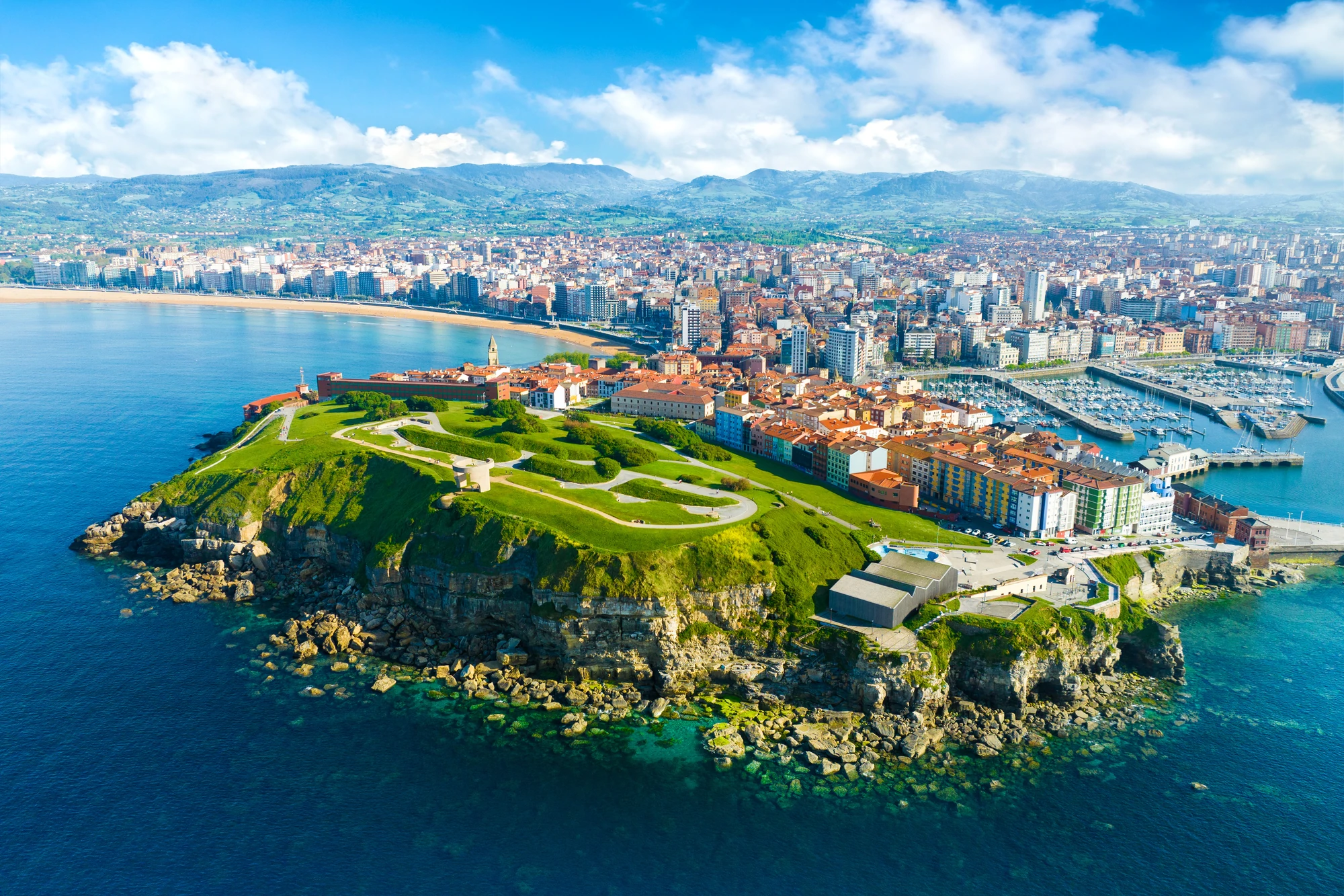 Gijon Luftaufnahme einer grünen Landzunge mit befestigten Wegen, umgeben von Meer, im Hintergrund eine Stadt mit vielen Gebäuden und einem Hafen.