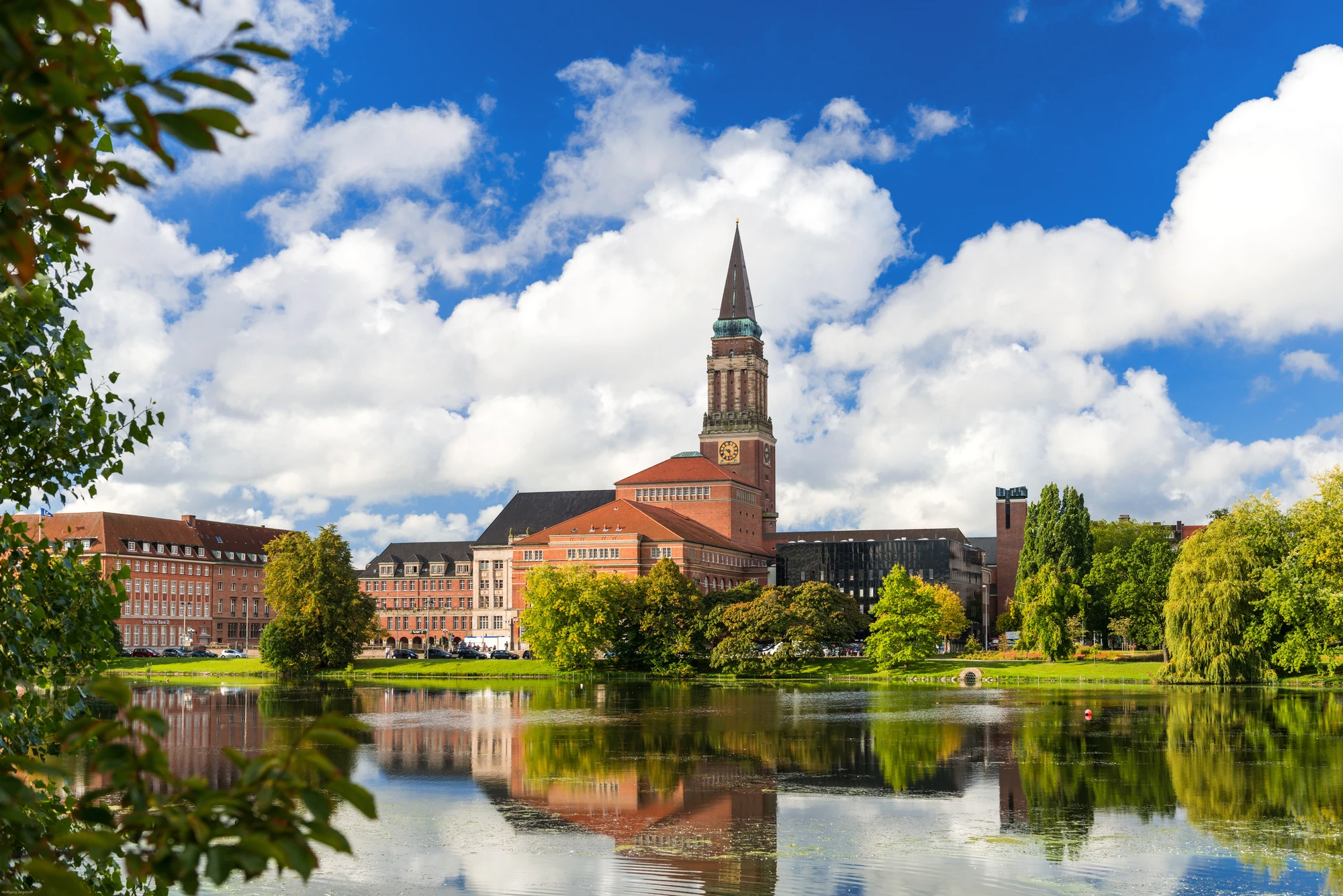 Kiel_ Stadtansicht mit einem historischen Gebäude mit Turm, umgeben von Bäumen und einem ruhigen See, unter einem blauen Himmel mit Wolken.