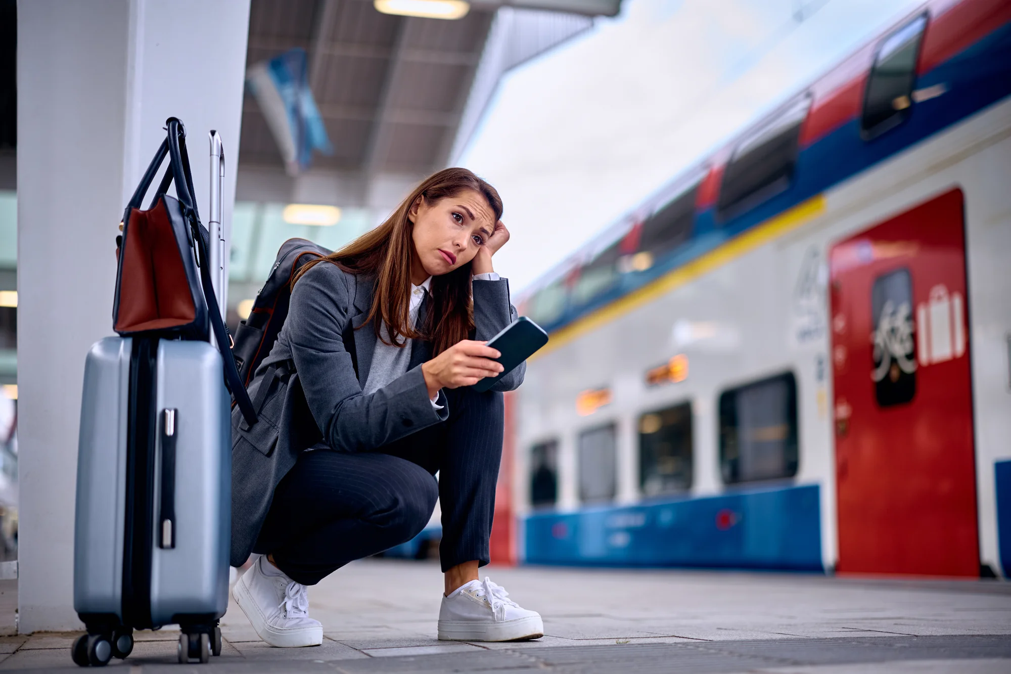 Eine Frau in Business-Kleidung sitzt nachdenklich und mit einem besorgten Ausdruck auf einem Bahnsteig, während sie auf ihr Smartphone schaut. Neben ihr steht ein Koffer, was die Situation einer möglichen Reiseverzögerung oder eines Warteproblems symbolisiert. Im Hintergrund ist ein Zug zu sehen, der den Kontext einer Bahnfahrt unterstreicht.