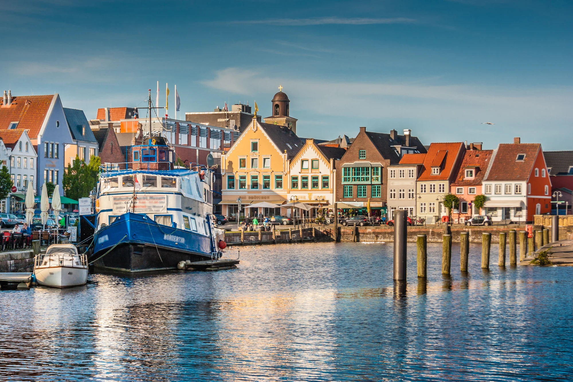 Blick auf einen Hafen mit einem blauen Schiff und mehreren Gebäuden in verschiedenen Farben im Hintergrund. Wasser spiegelt die Szenerie wider. _ Kiel
