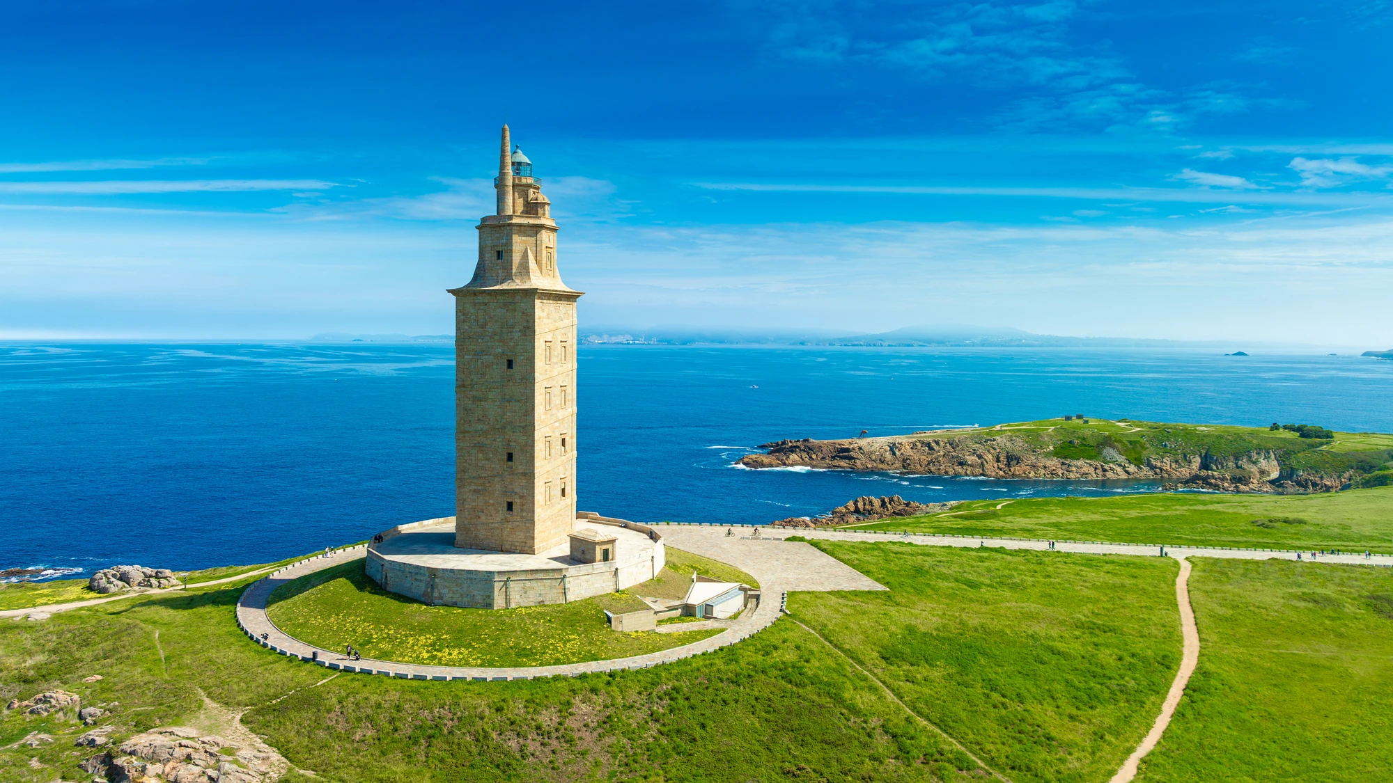 Der Herkulesturm, ein steinerner Leuchtturm, steht auf einer grünen Landzunge am Meer. Der Himmel ist klar und blau, das Wasser ist ruhig.