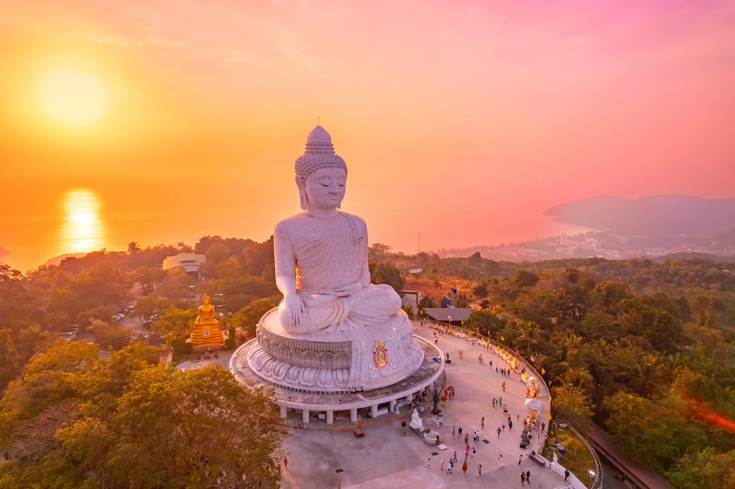 Phuket _ Große weiße Buddha-Statue auf einem Hügel mit Blick auf die Küste bei Sonnenuntergang, umgeben von Bäumen und Besuchern