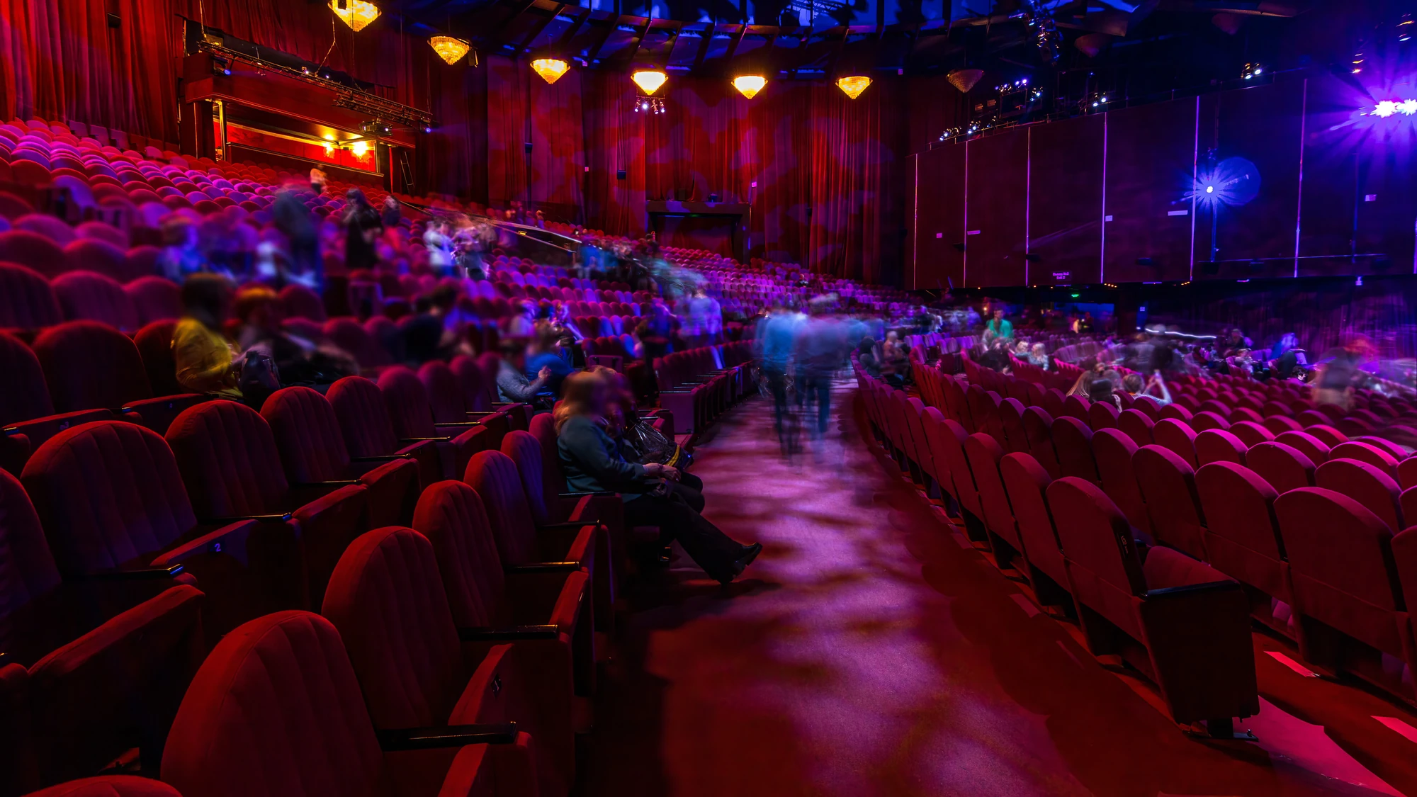 Theatersaal mit roten Sesseln und mehreren Zuschauern, einige Personen sind durch Bewegungsunschärfe verschwommen