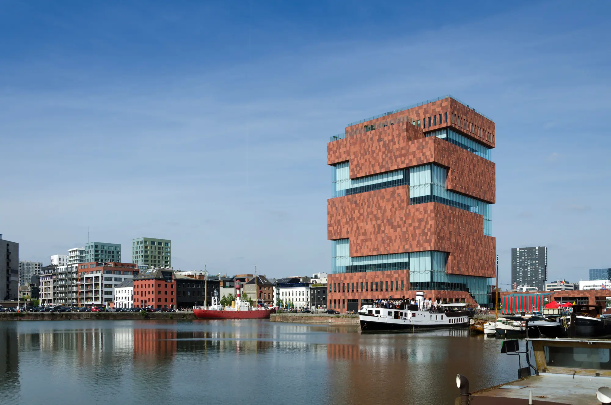 Das MAS-Museum in Antwerpen, ein markantes, gestapeltes Gebäude aus rotem Sandstein und Glas, am Hafen gelegen. Im Vordergrund liegen Schiffe auf ruhigem Wasser, der Himmel ist klar.