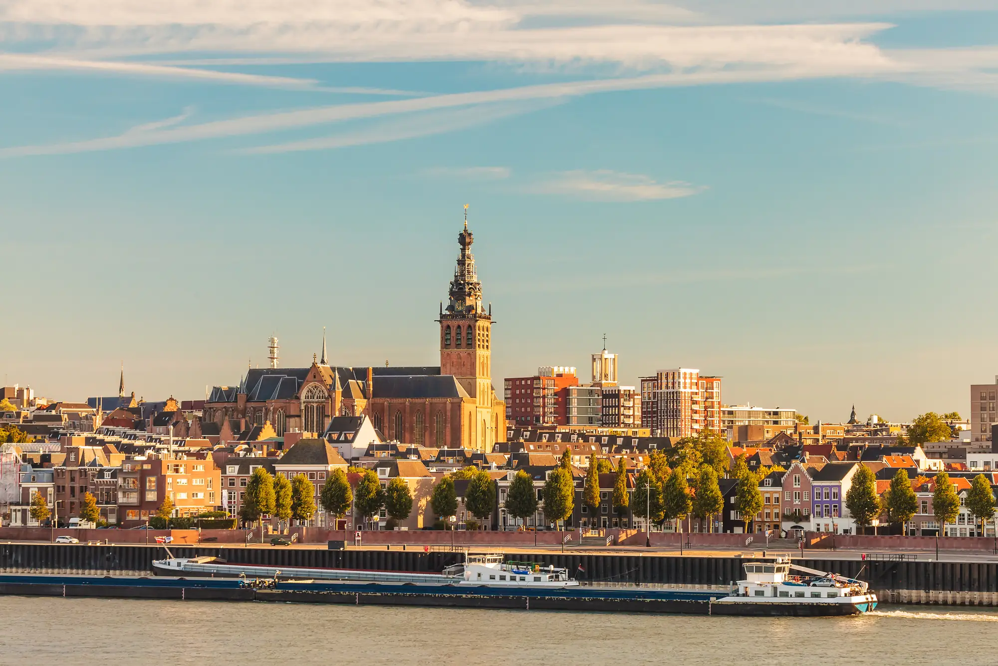 Stadtansicht mit Fluss im Vordergrund, einem langen Frachtschiff und einer großen Kirche mit hohem Turm in der MitteStadtansicht mit Fluss im Vordergrund, einem langen Frachtschiff und einer großen Kirche mit hohem Turm in der Mitte