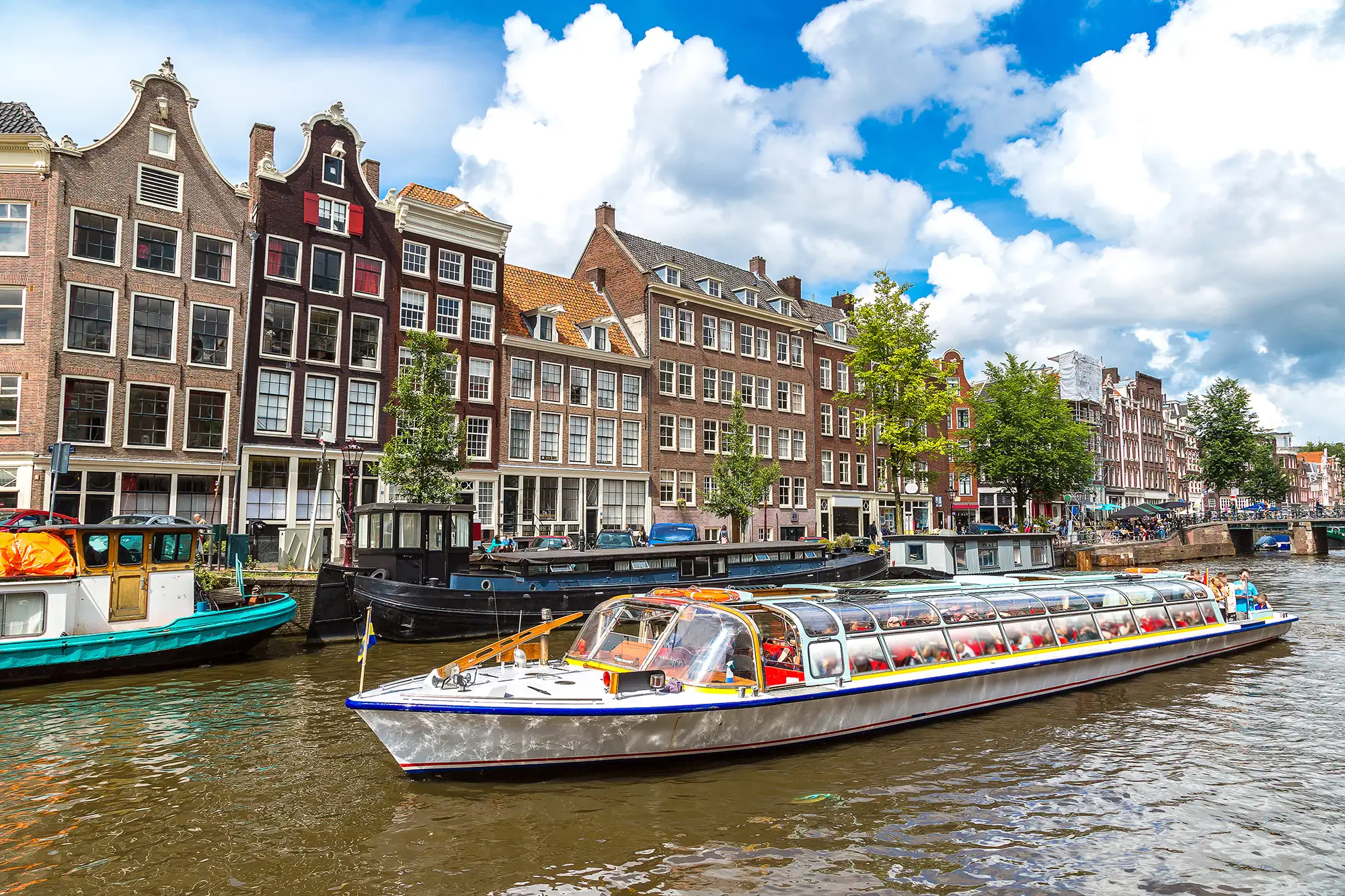 Boot auf einer Gracht in Amsterdam bei leicht sonnigem Wetter, das Wasser glitzert im Sonnenlicht und reflektiert die umgebenden Häuser und Bäume.