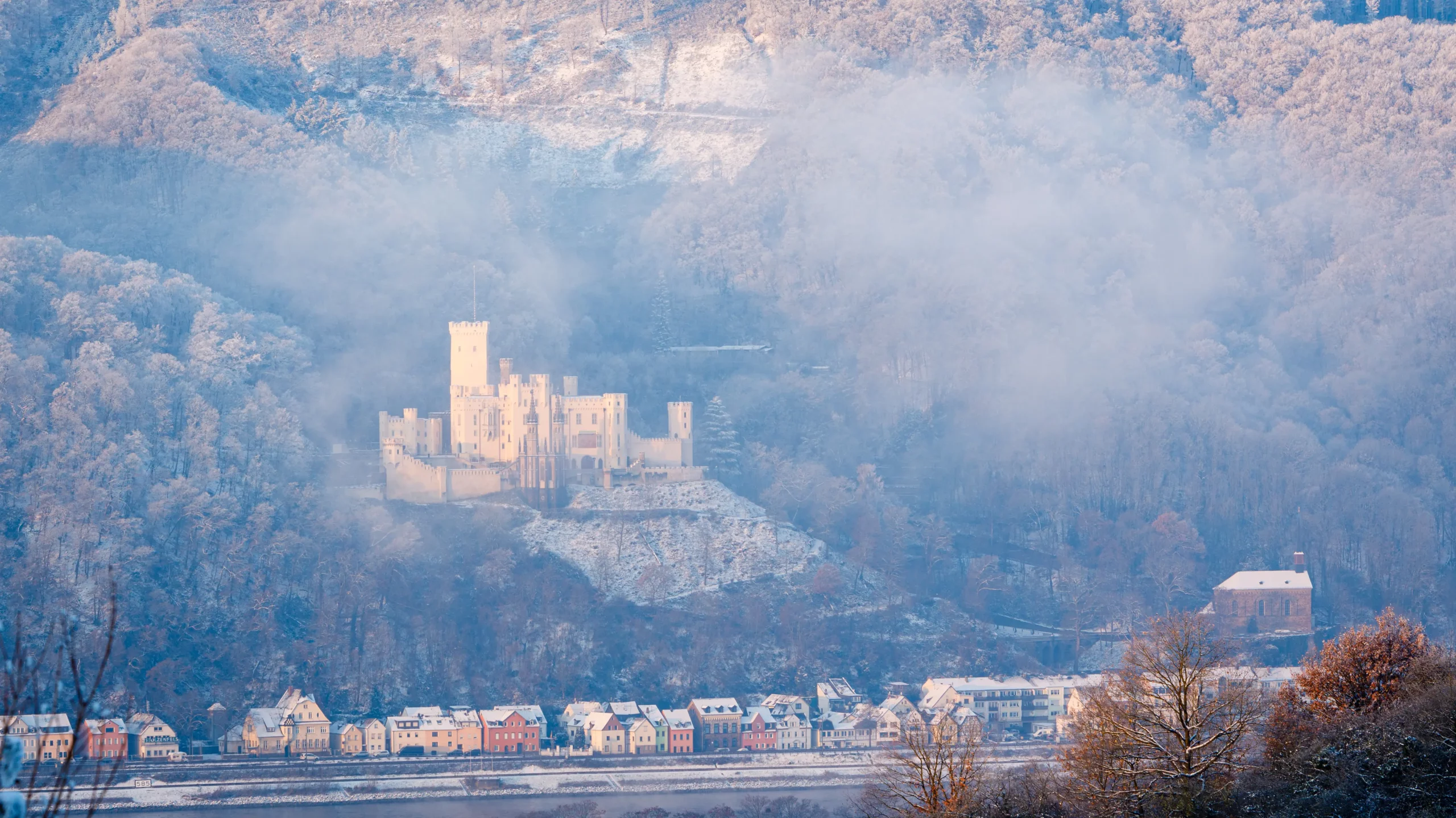 Schloss auf einem bewaldeten Hügel mit Nebel, darunter eine Reihe bunter Häuser am Fuß des Hügels Winteraufnahme von Schloss Stolzenfels in Koblenz am Rhein, Rheinland-Pfalz, Deutschland, Europa.